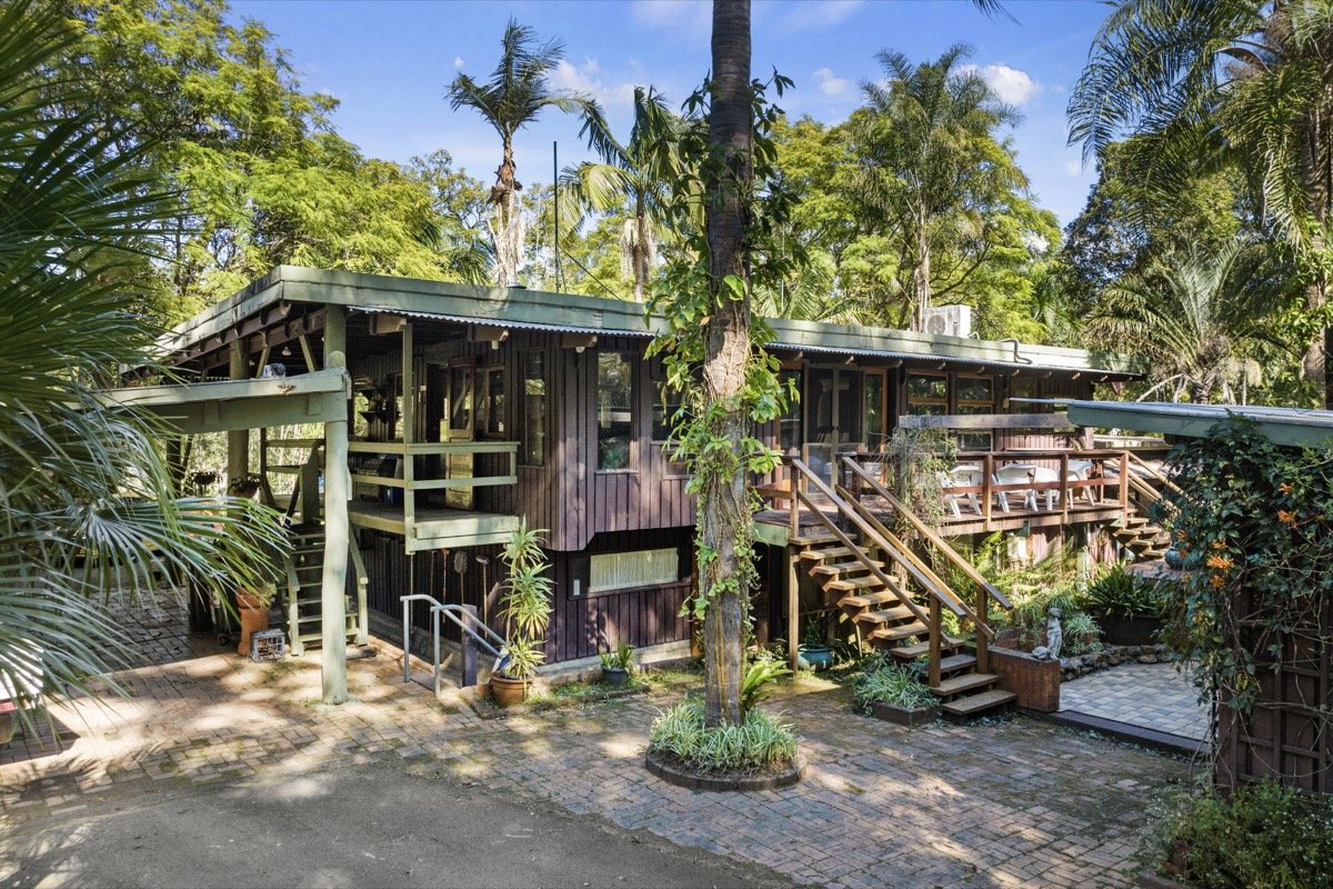 Wooden house on stilts surrounded by lush green tropical trees and plants, with an outdoor patio, stairs, and a brick walkway.