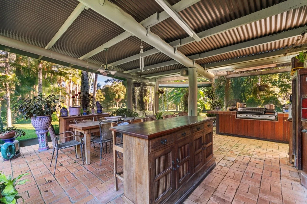 A screened-in outdoor kitchen and dining area with brick flooring, a wooden island, a large table with chairs, a grill station, and potted plants, surrounded by trees and greenery.