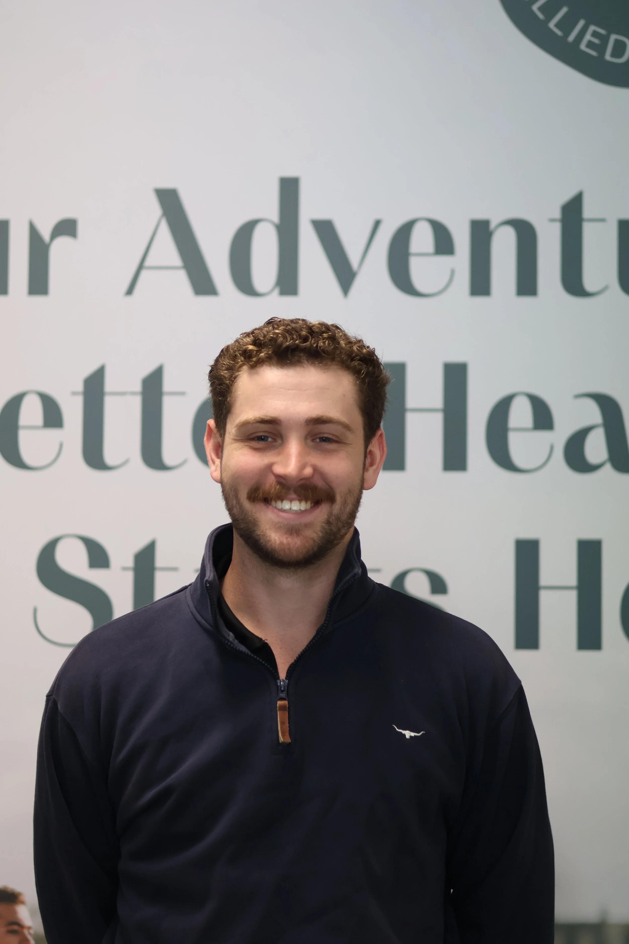 Young man with curly brown hair and a beard smiling, wearing a dark zip-up sports jacket, standing in front of a large sign with the text 'Our Adventure' and 'St' visible.