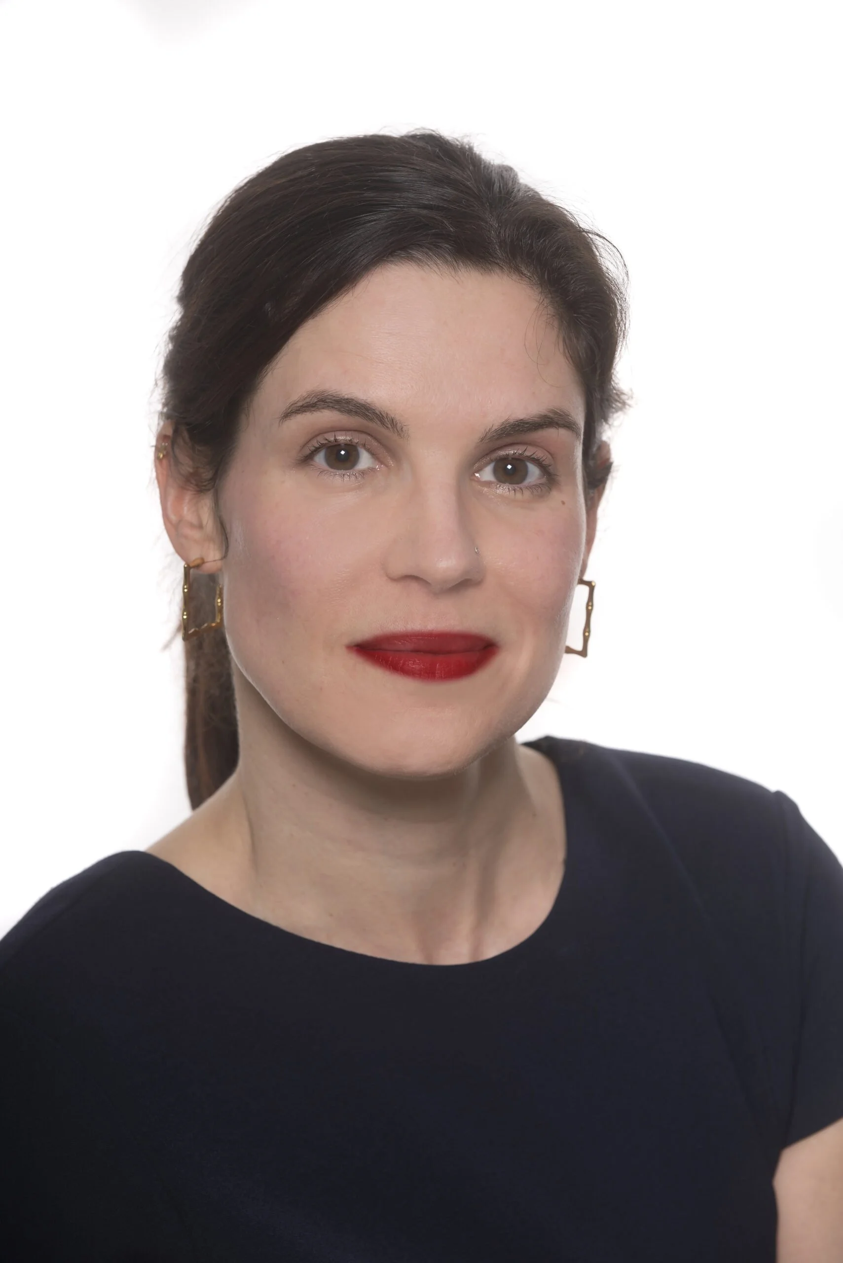 Close-up portrait of a woman with dark brown hair, fair skin, red lipstick, and gold geometric earrings, wearing a black top, against a white background.