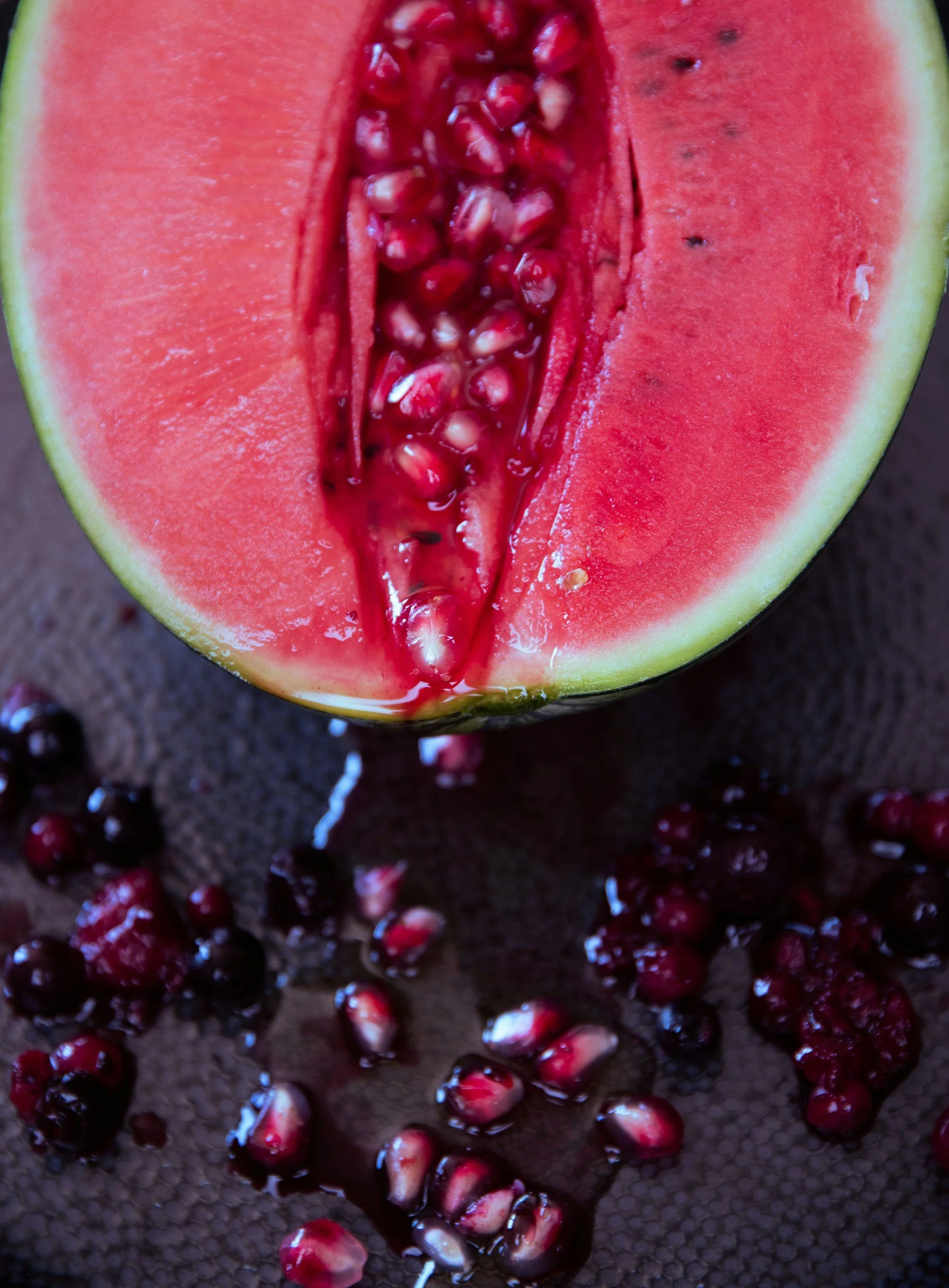 A close-up of a halved watermelon with bright red flesh and black seeds, surrounded by scattered pomegranate seeds on a dark textured surface.