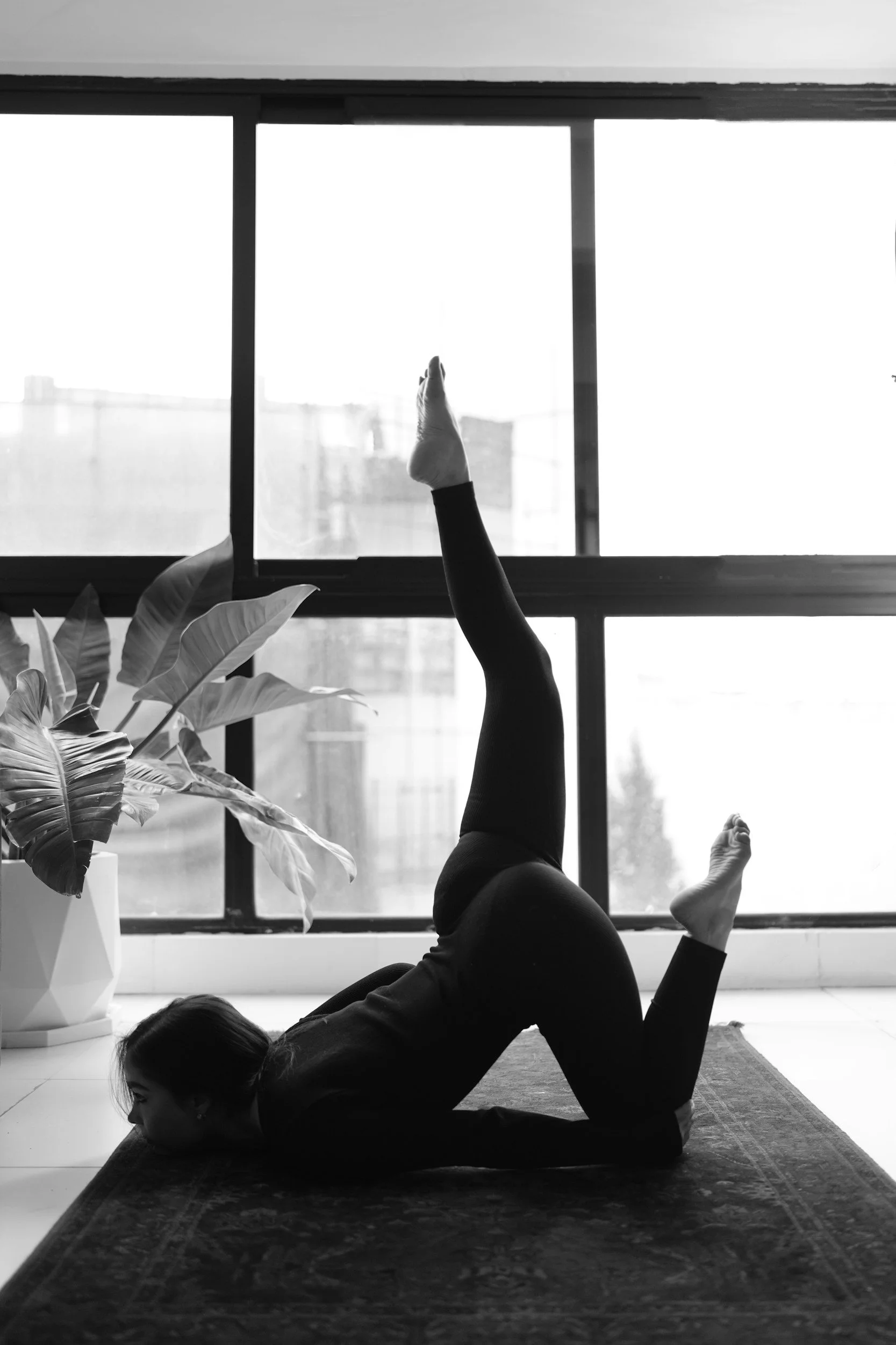 A woman doing yoga indoors, lying on her stomach on a mat with one leg raised vertically, near a large window with cityscape view, with a planter plant beside her.