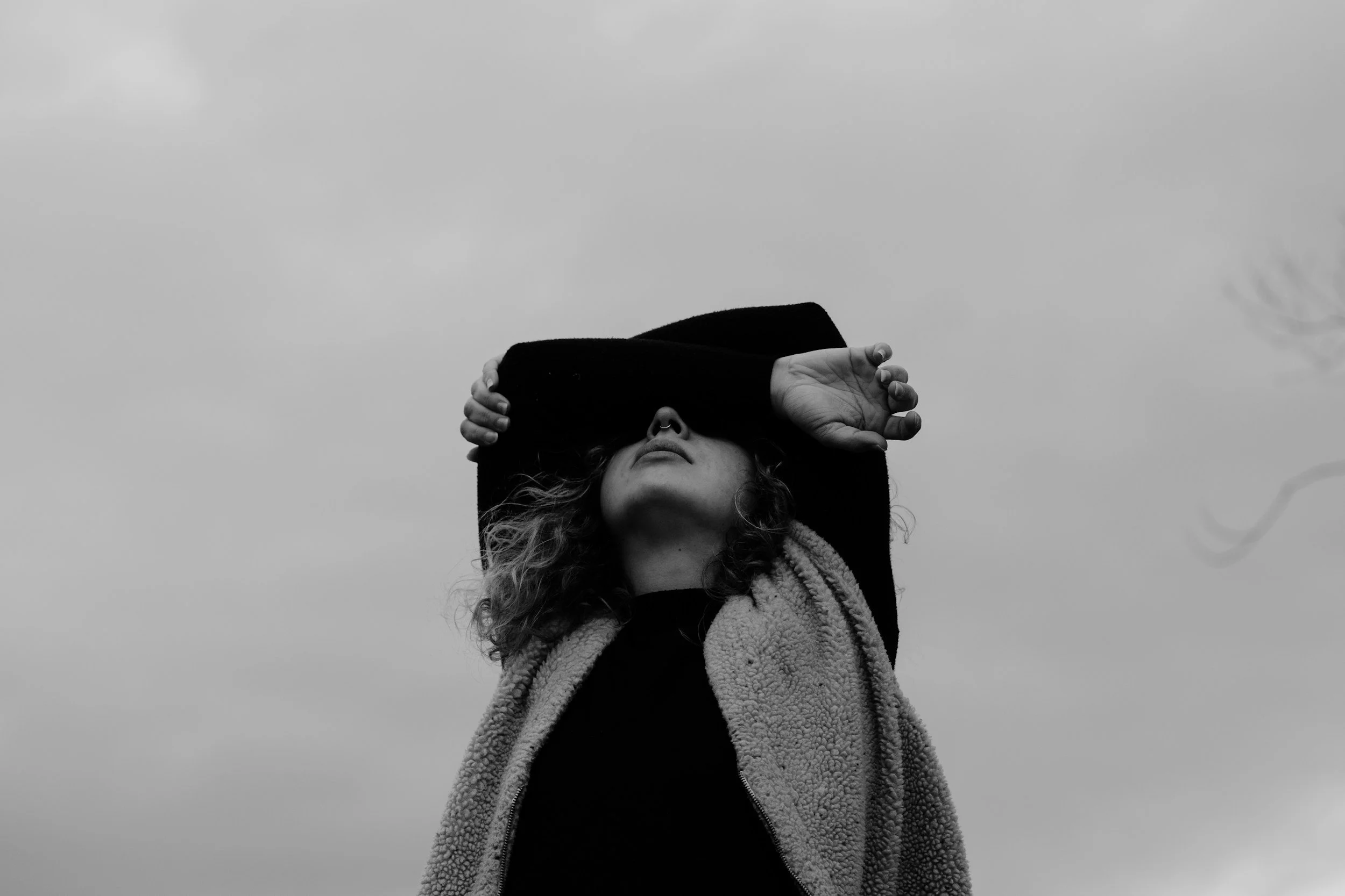 A black and white photo of a woman with curly hair, wearing a fleece jacket, covering her eyes with her arm against a cloudy sky.