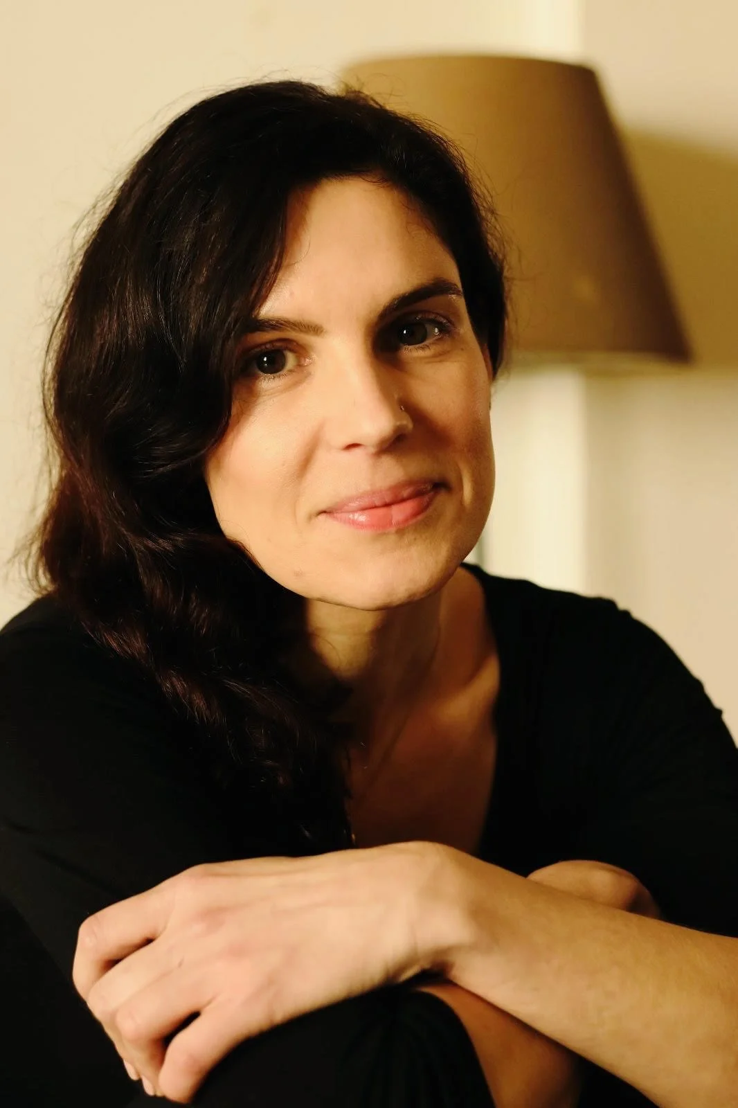 A woman with dark hair and a nose piercing, wearing a black top, sitting with her arms crossed on a table, in a warmly lit room.