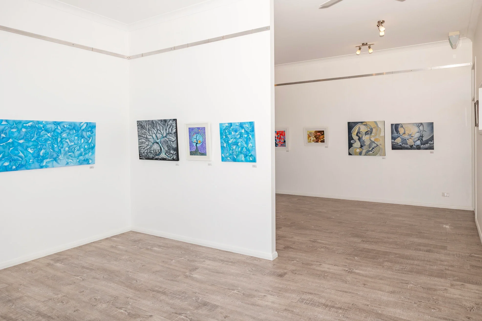An art gallery with white walls and wooden flooring displaying colorful abstract paintings. The left side shows blue and gray artwork, and the right side features paintings with warm tones. Ceiling spotlights illuminate the artworks.