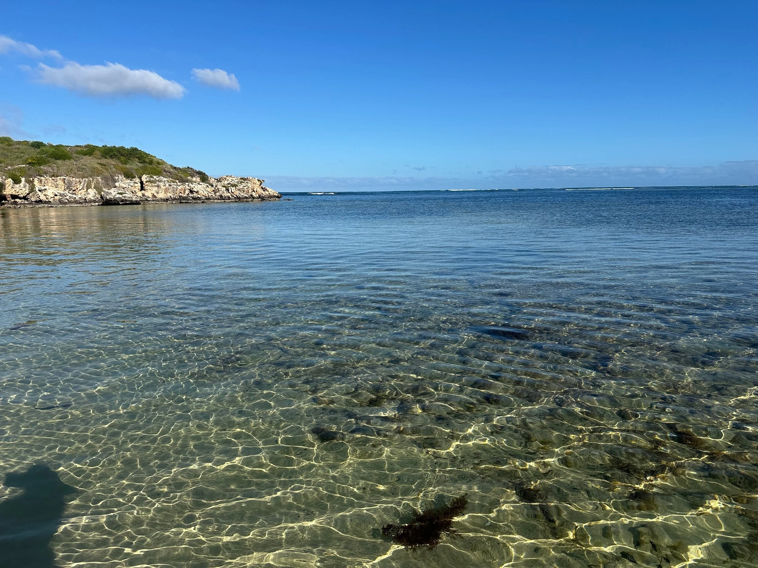 Clear shallow water near a rocky coastline with a hillside on the left, under a bright blue sky with some clouds.