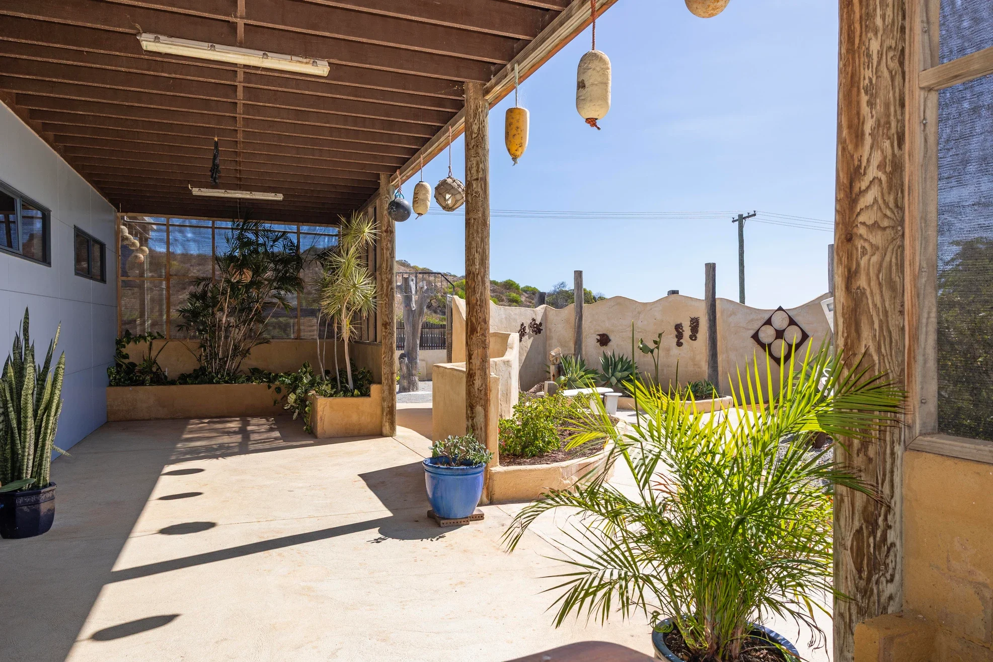 A covered outdoor patio area with potted plants, hanging decorative buoys, and a stucco wall in the background under a clear blue sky.