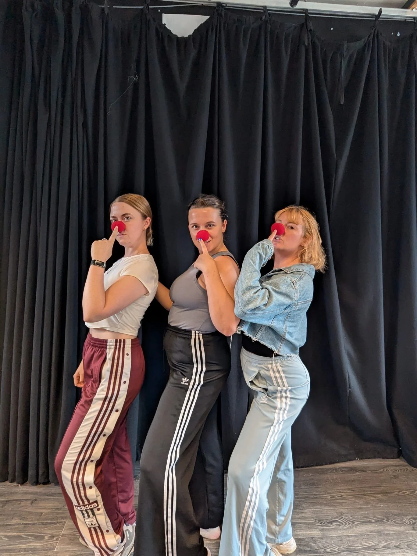 Three women standing side by side in front of a black curtain, each with a red clown nose, striking playful poses.