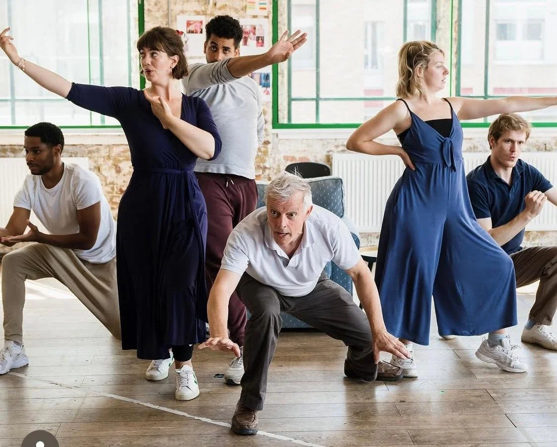 Group of five people, four young adults and one older man, practicing or rehearsing a dance or theater routine in a studio with large windows, hardwood floors, and exposed brick walls.