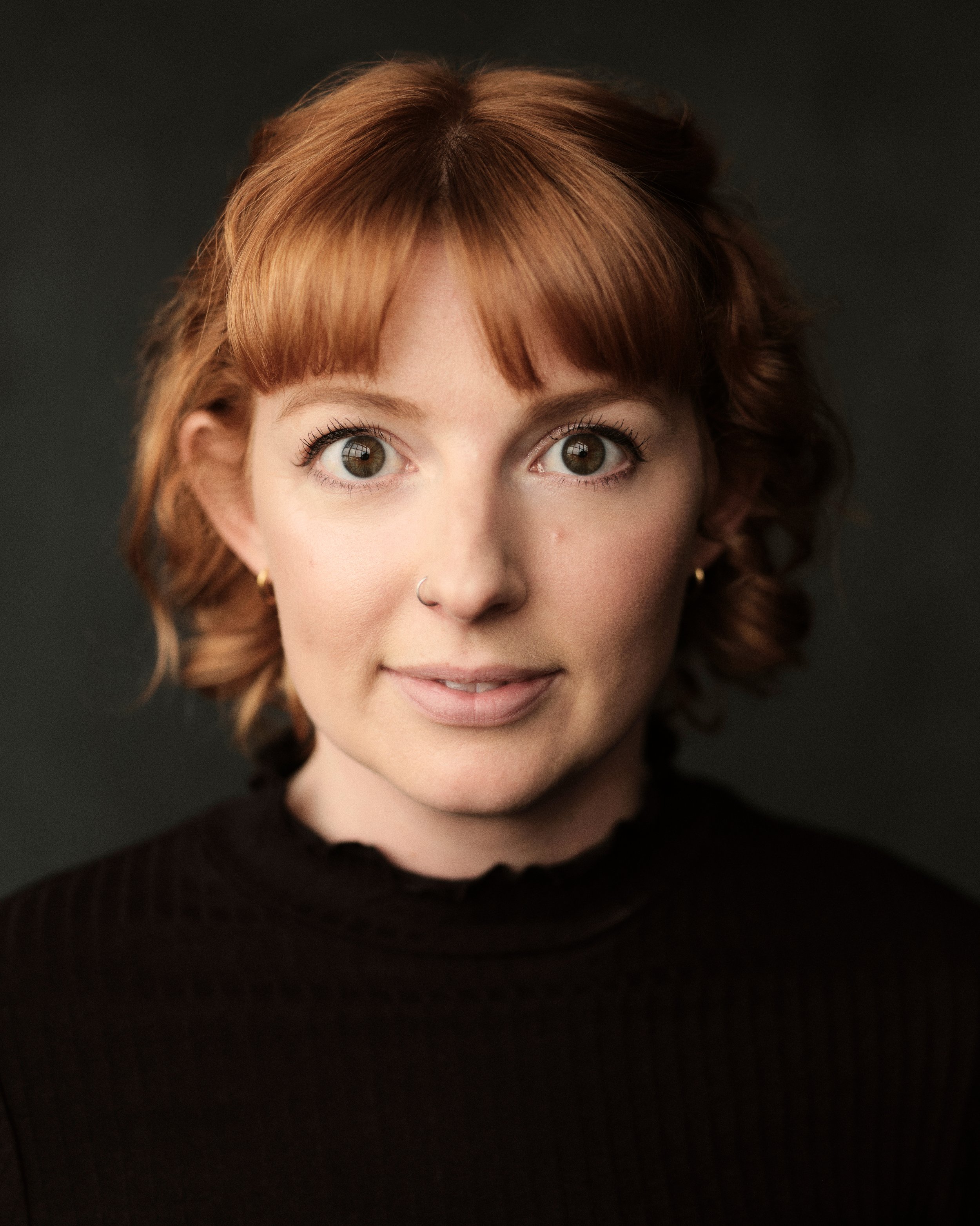 Close-up of a woman with short red hair, brown eyes, nose piercing, and wearing a black top, against a dark background.