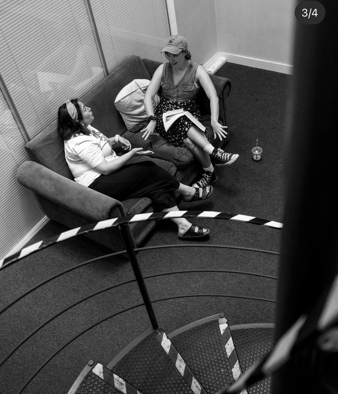 Two women are sitting on a couch, engaged in a conversation in a room with blinds and a corner wall. One woman, wearing a hat, a sleeveless denim vest, and a floral skirt, is speaking animatedly. The other woman, wearing glasses, a headband, a white top, and dark pants, is listening. There is a small drink on the floor nearby and a striped barrier in the foreground, indicating the photo was taken from a staircase or elevated position.