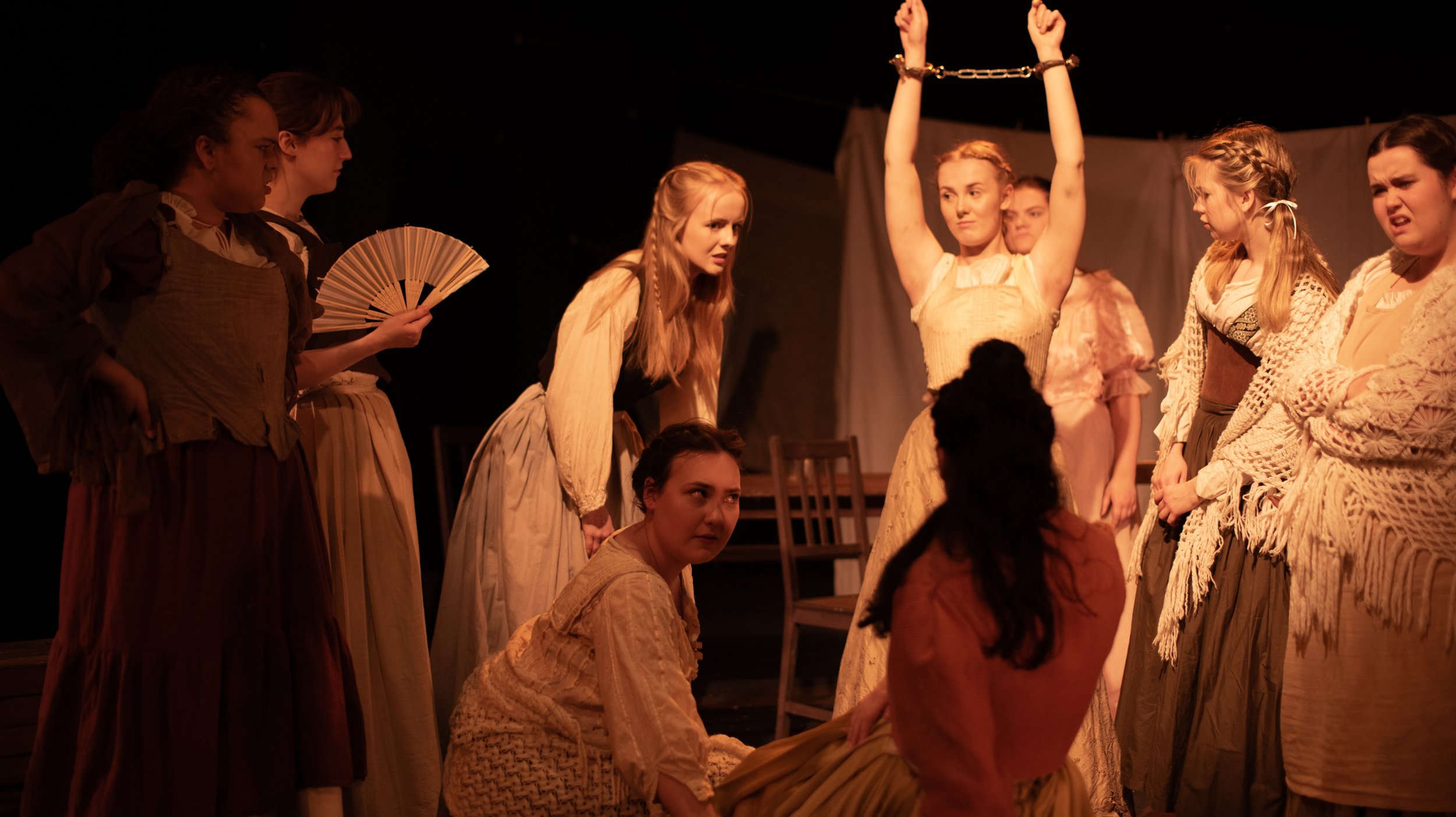 The scene shows a theatrical performance with several women dressed in vintage or historical costumes. One woman is handcuffed and raising her hands, while others are standing or sitting around her, engaging in intense expressions and interactions.