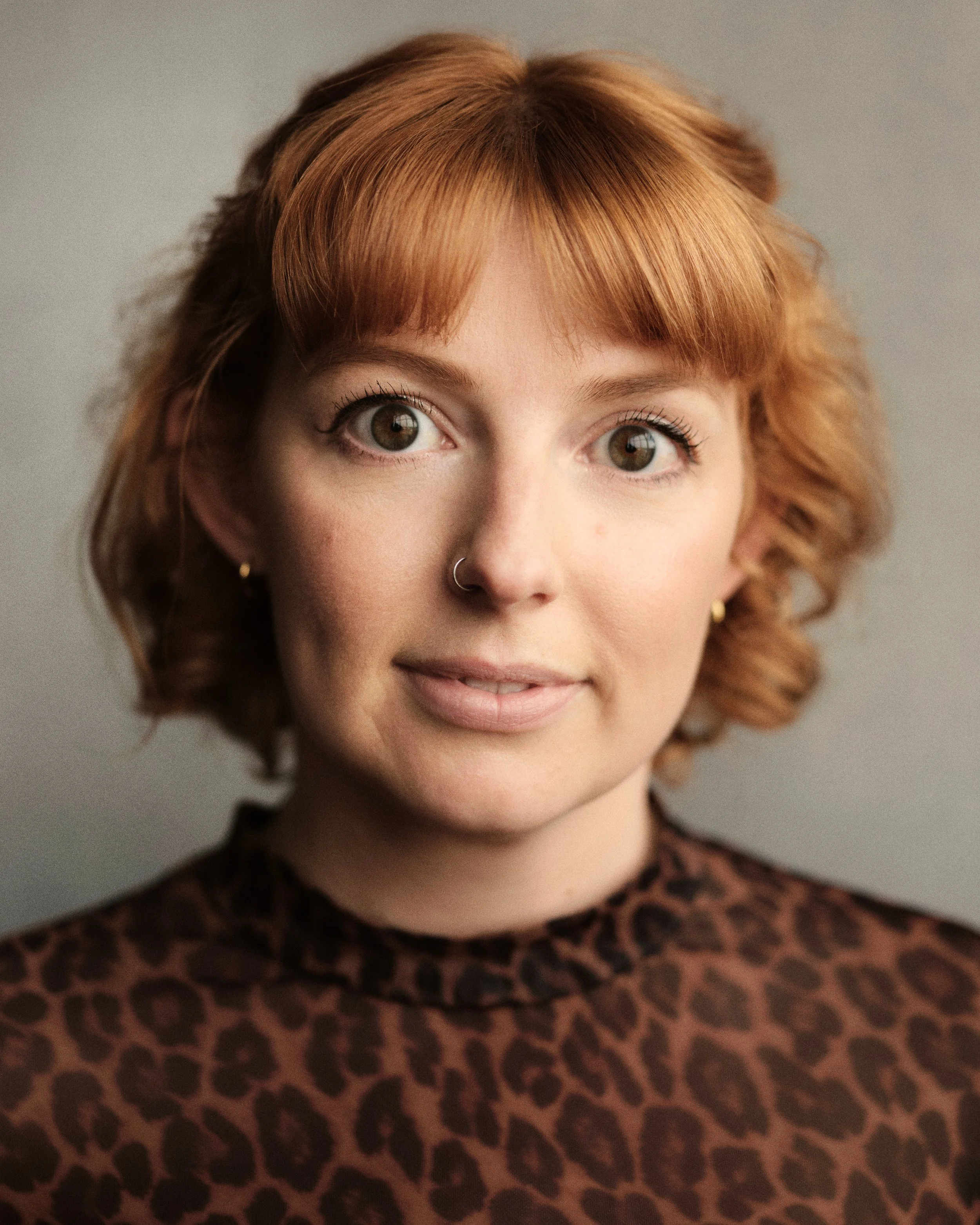 Close-up of a woman with short, wavy auburn hair and hazel eyes, smiling gently, wearing a leopard print top.
