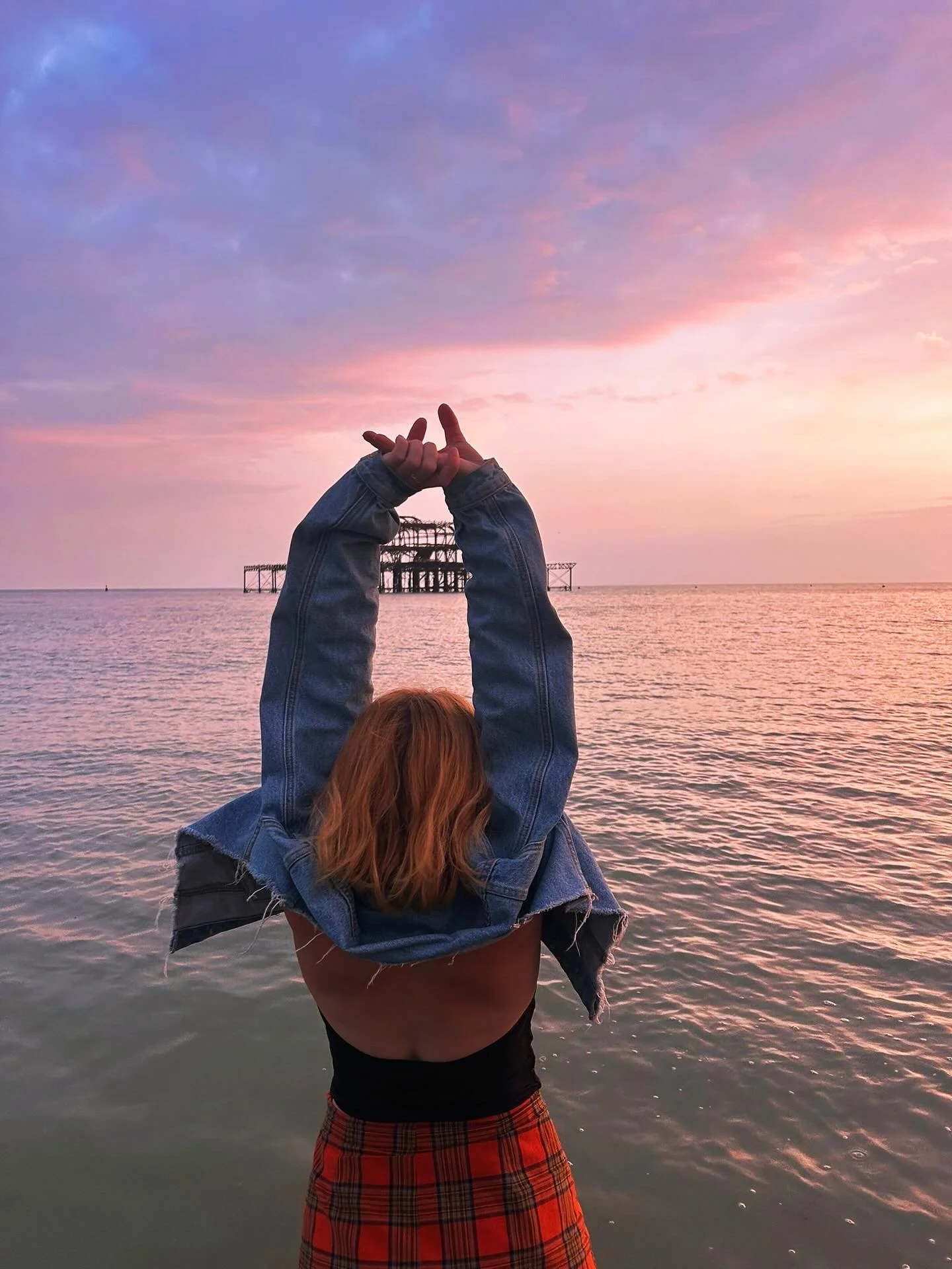 Person with red hair and plaid skirt holding another person with a denim jacket on their shoulders, standing by the water with a sunset sky and an old pier in the background.