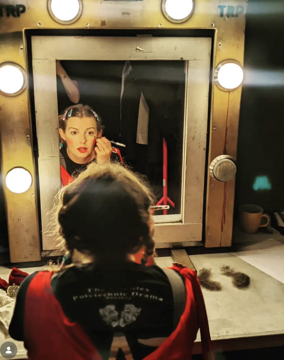 A woman is looking into a mirror while applying makeup, with her reflection visible. The scene appears to be a backstage or dressing room setting with bright lights around the mirror. A person with braided hair, wearing a black and red shirt with a clown face and the words 'Theatre' visible, is sitting in front of the mirror. The countertop has makeup items, hair strands, and a mug on it.