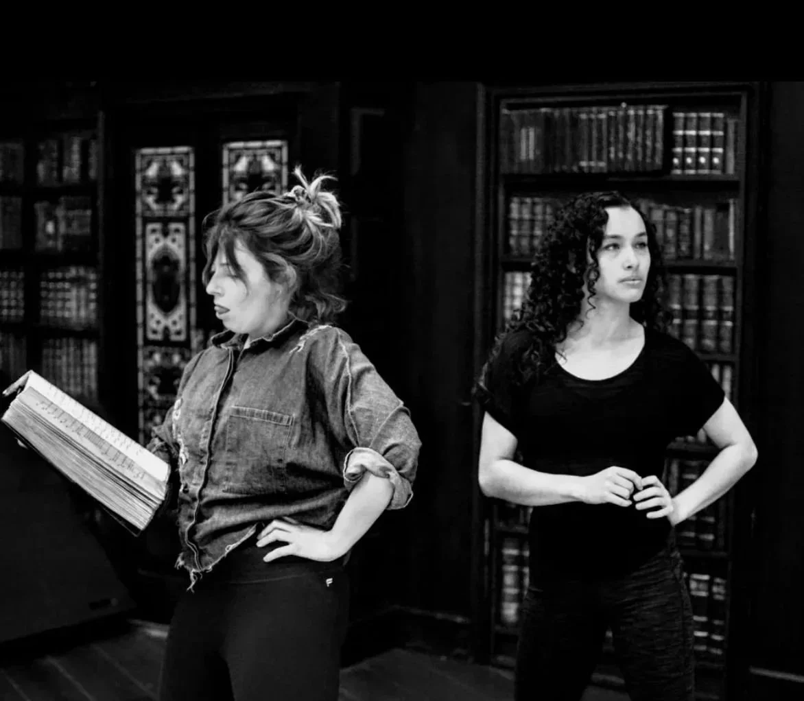 Two women standing in front of bookshelves in a library, one reading a book and the other looking away with hands on hips, black and white photo.