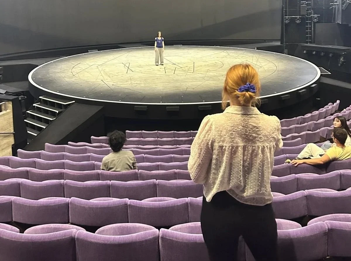 A woman with red hair in a bun, wearing a beige blouse, observes a stage with a woman standing on it, equipped with purple seats and stairs leading up to the stage, likely in a theater or performance space.