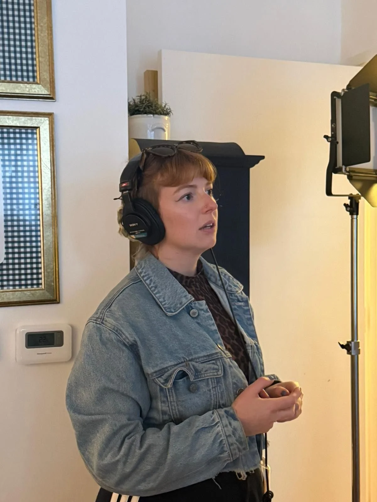 A woman with short red hair, wearing a denim jacket, black top, large headphones, and glasses on her head, stands in front of a studio light, holding a small remote or microphone in her hands, indoors.