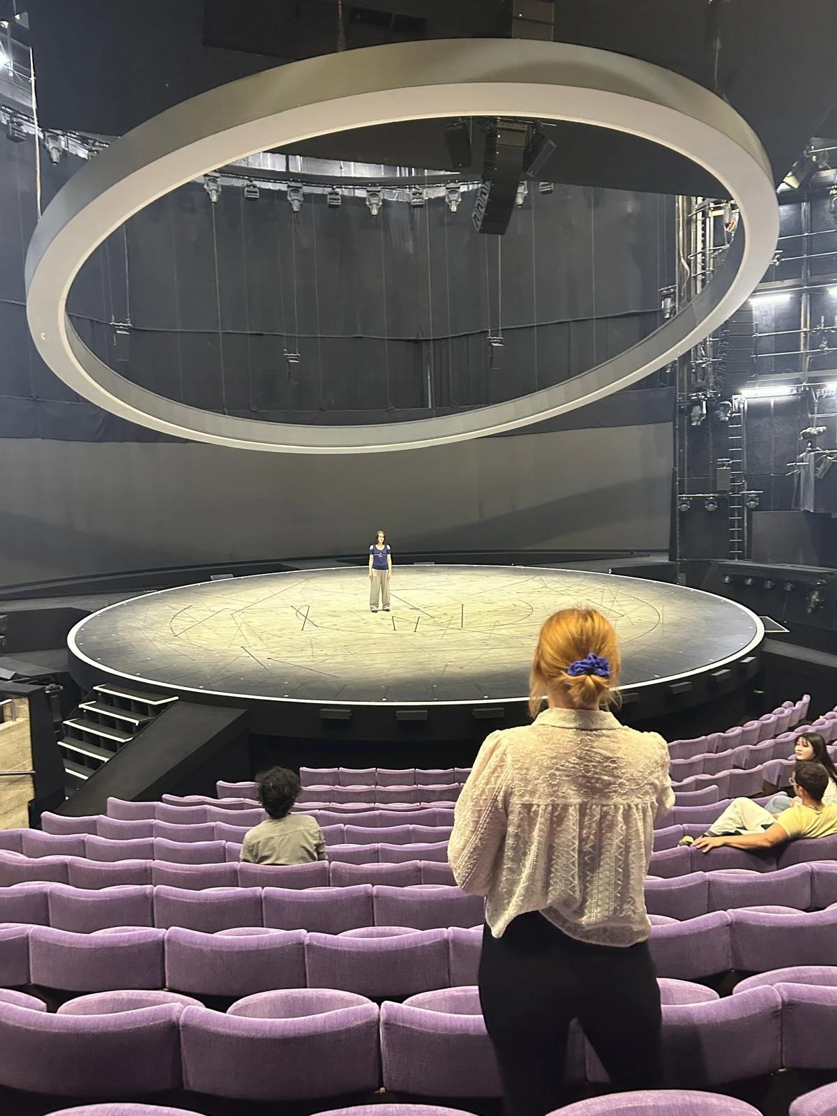 The image shows an empty theater with purple seats. A woman with red hair tied back is standing near the seats, observing a person on a stage. The stage is circular with a person standing at the center, and a large oval structure is hanging above the stage, part of the theater's modern stage design.
