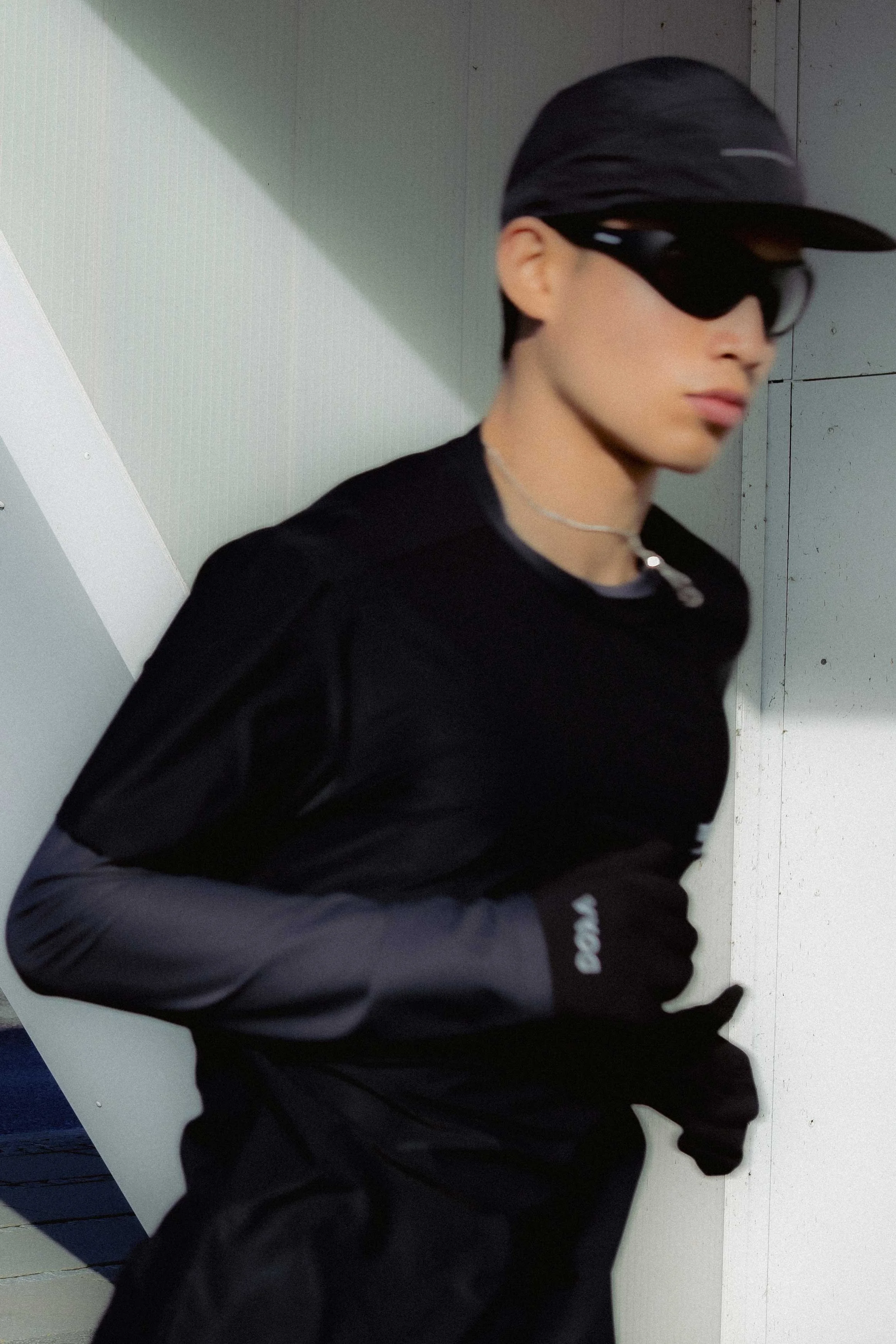 A person wearing a black cap, black sunglasses, a black shirt, and black gloves, standing against a wall with a shadow cast on it.