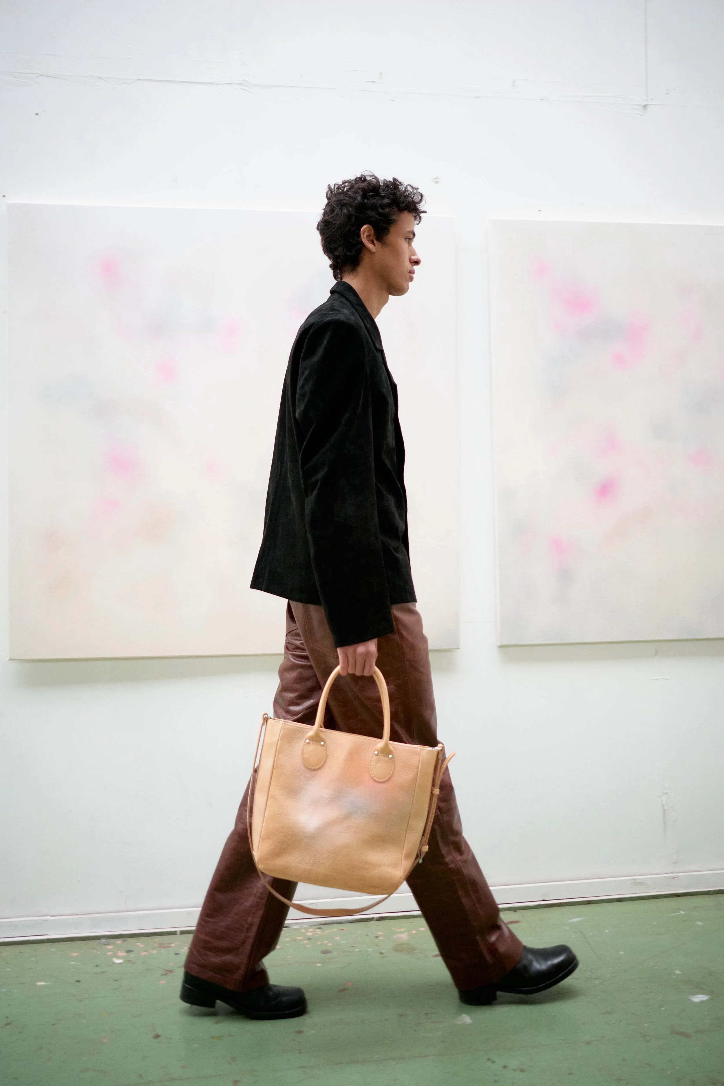 A person with short curly hair walking past white walls with pink marks, carrying a large tan handbag, wearing brown pants and black shoes.