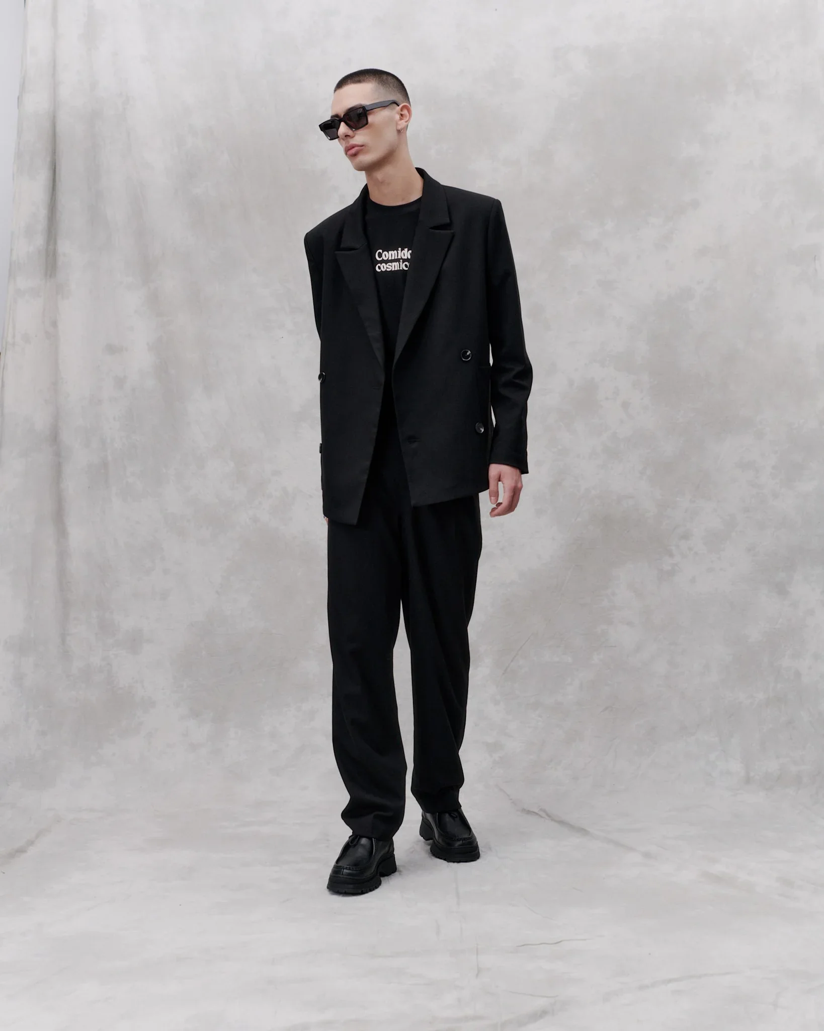 A guy, wearing black sunglasses, a black double breasted blazer and trousers.