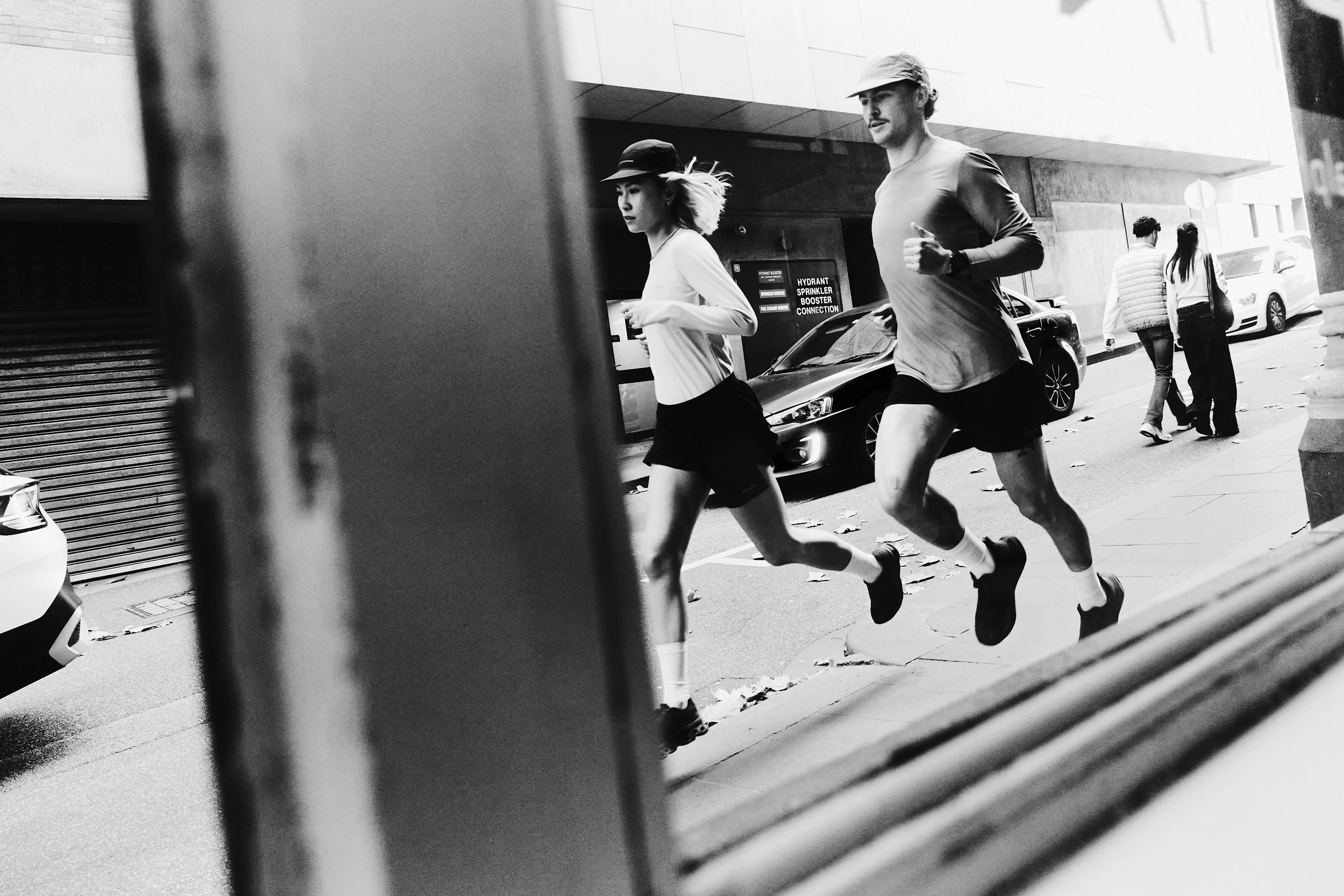 Two runners jogging on a city sidewalk, captured through a vertical opening in a fence or barrier, with parked cars and pedestrians in the background.