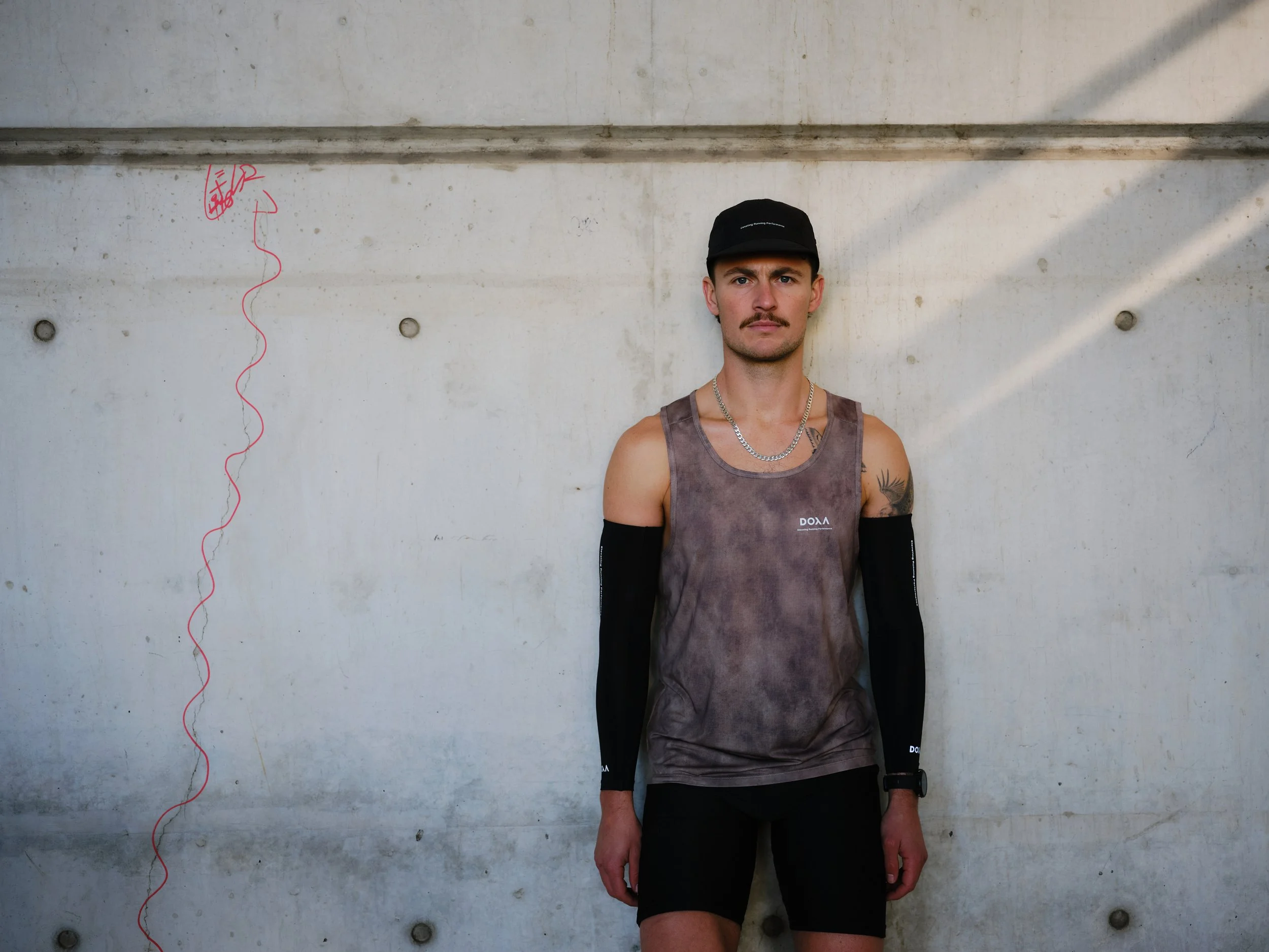 A man standing against a concrete wall, wearing a black cap, sleeveless shirt, and black sleeves, with tattoos on his arm and a chain necklace.