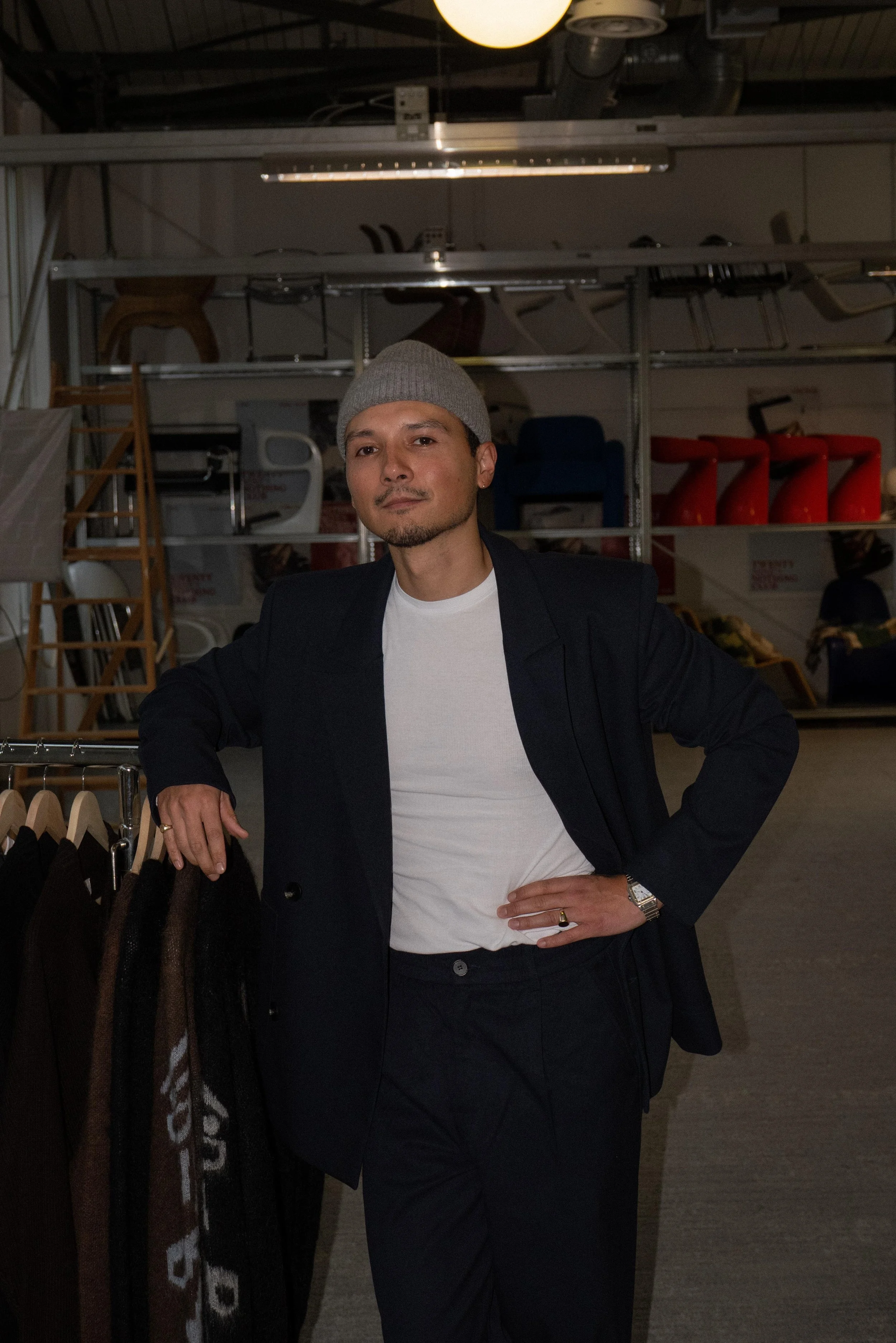 A man wearing a grey beanie, white t-shirt, black blazer, and black pants, standing with one hand on his hip and the other resting on a clothing rack, inside a warehouse store, with shelves of chairs and red objects in the background.