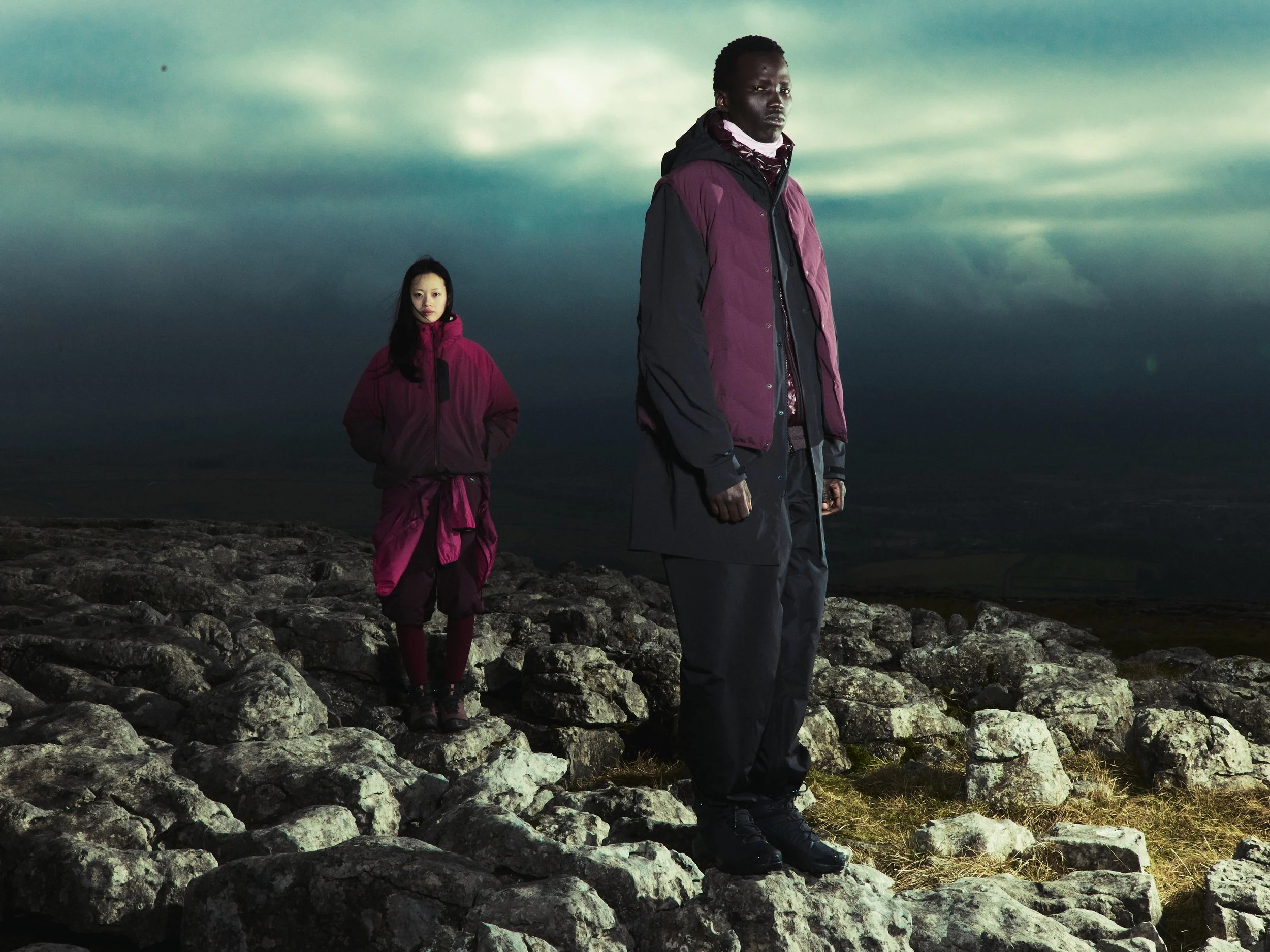 Two individuals dressed in dark outdoor clothing standing on rocks, with cloudy sky in the background, during dusk or dawn.