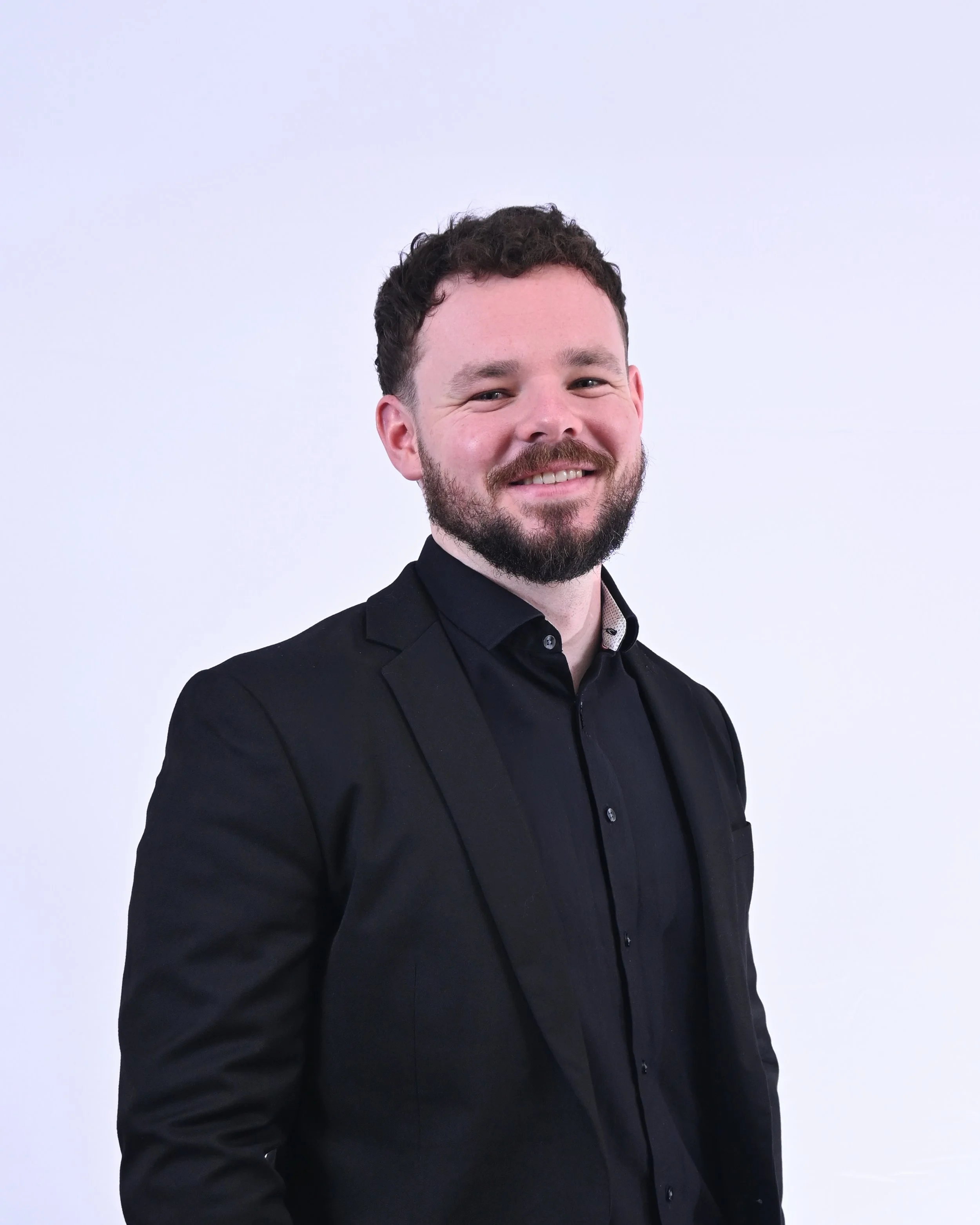 A man with curly dark hair and a beard wearing a black suit and black shirt, smiling against a plain white background.