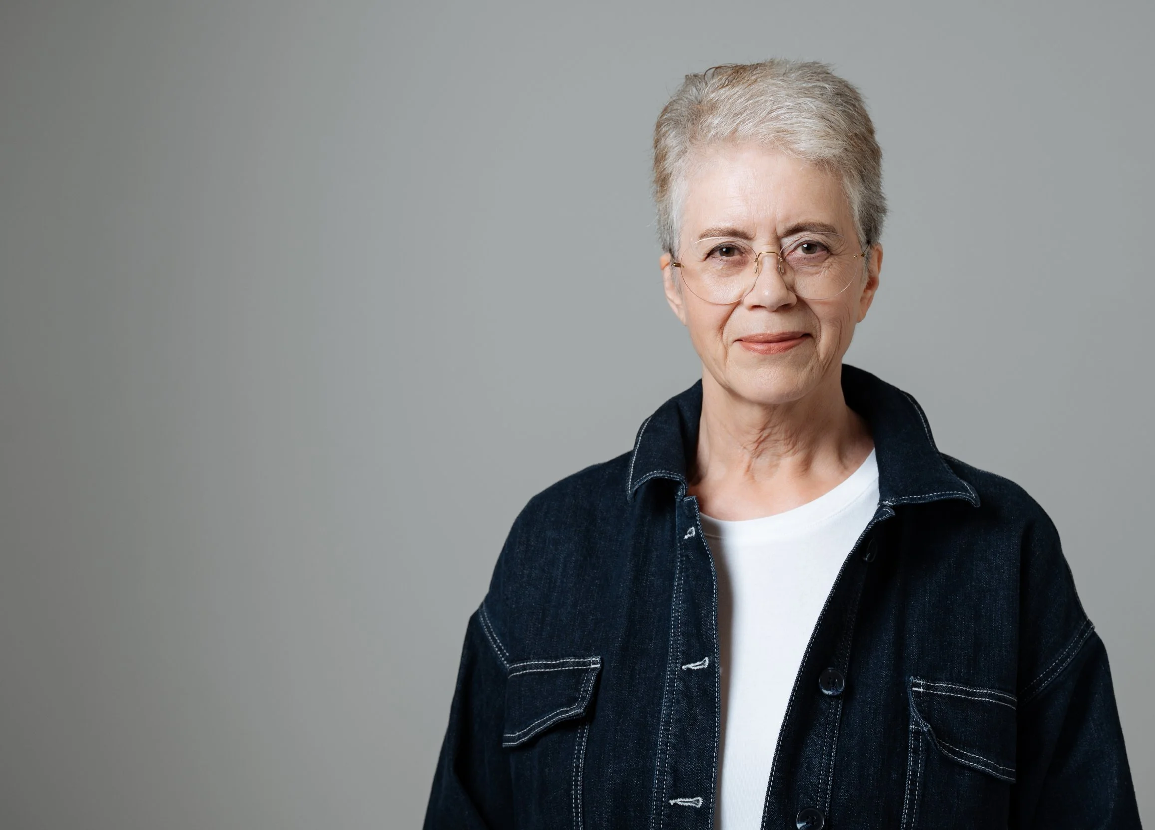 Ältere Frau mit kurzen grauen Haaren, Brille, weißem T-Shirt und dunkler Jeansjacke vor grauem Hintergrund.