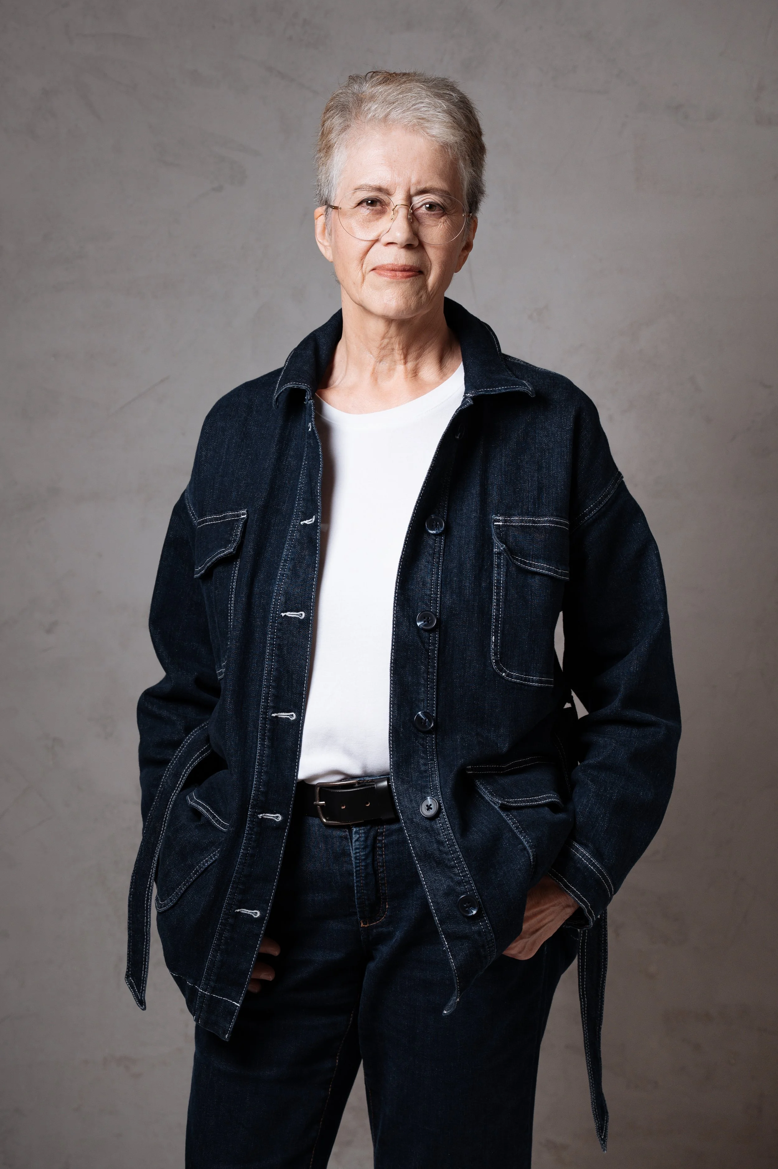 Porträt einer älteren Frau mit kurzen grauen Haaren, die eine schwarze Brille trägt, in einem dunkelblauen Jeansjacke, weißen T-Shirt und dunklen Hosen, vor grauem Hintergrund.