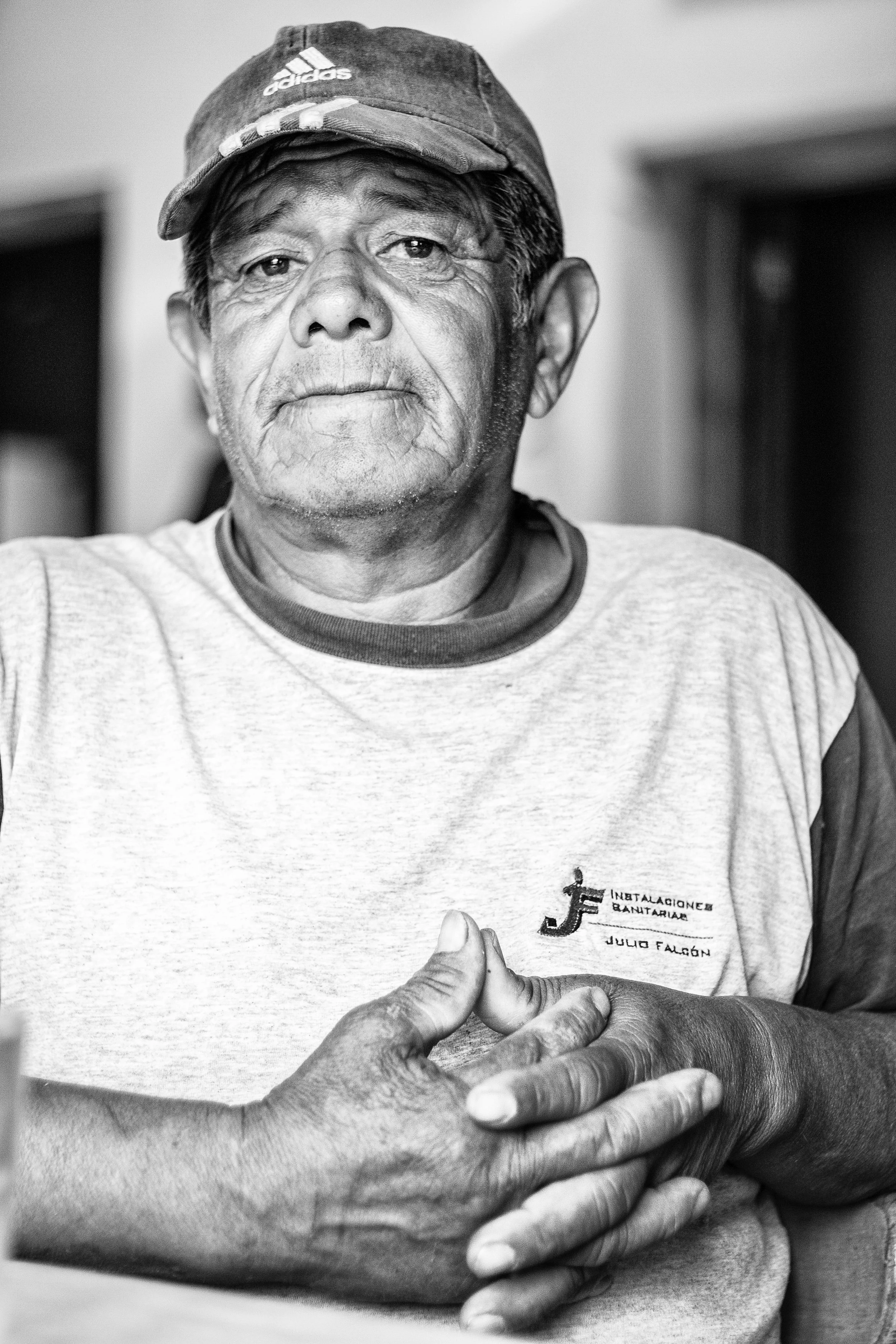 A black and white photo of an elderly man with short hair, wearing a baseball cap and a t-shirt. The man is giving a thumbs-up with his right hand, and his left arm is crossed over his chest.