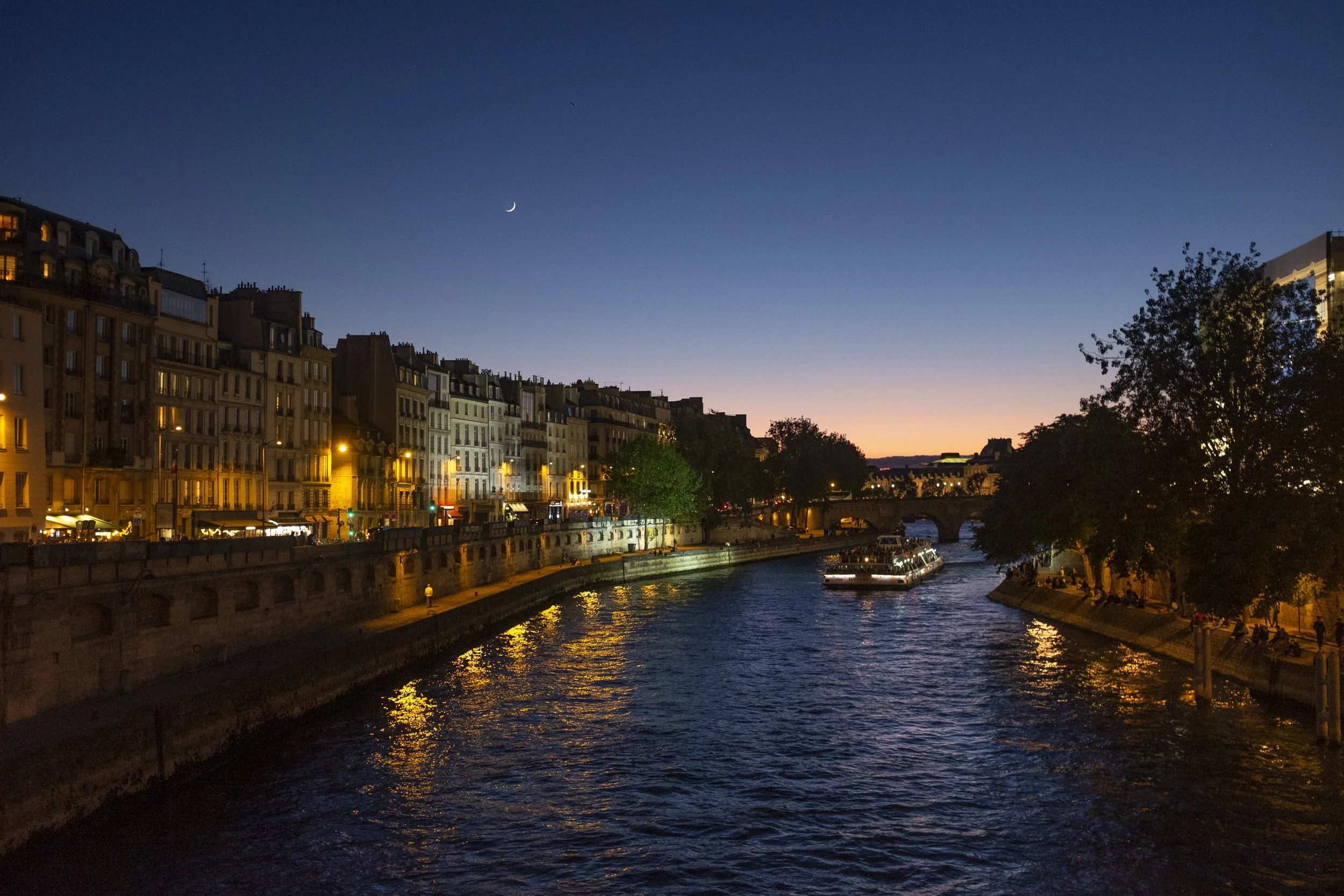 A cityscape at dusk along a river, with buildings illuminated by streetlights, a boat on the river, and a crescent moon in the clear sky.