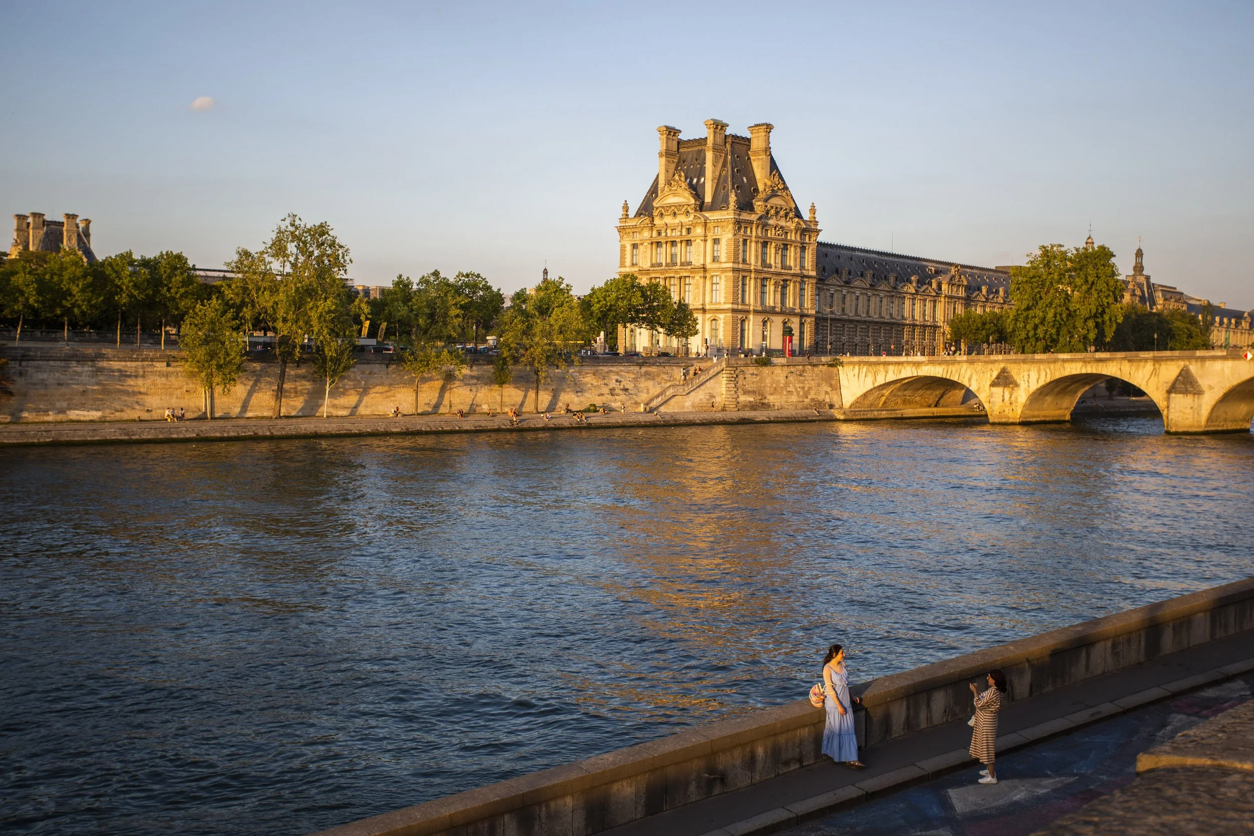 Two women stand on a riverbank at sunset, with historic buildings and trees across the water.