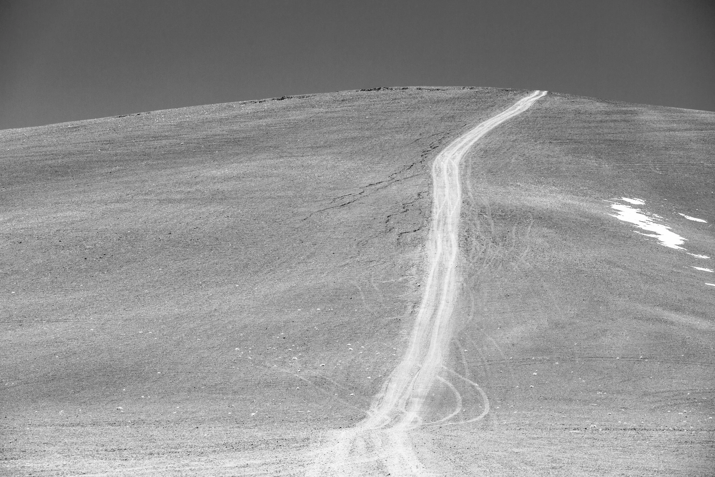 A black-and-white photograph of a barren, sloping landscape with a narrow, winding dirt trail running up the hill. The sky above is cloudless and dark.