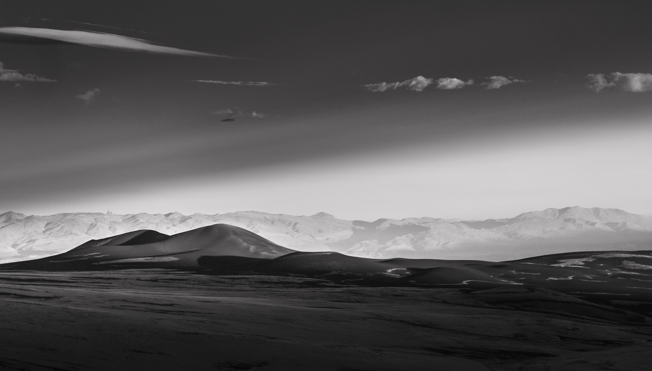 A black-and-white landscape featuring rolling hills and distant snow-capped mountains, with a sky filled with clouds and streaks of cloud formations overhead.