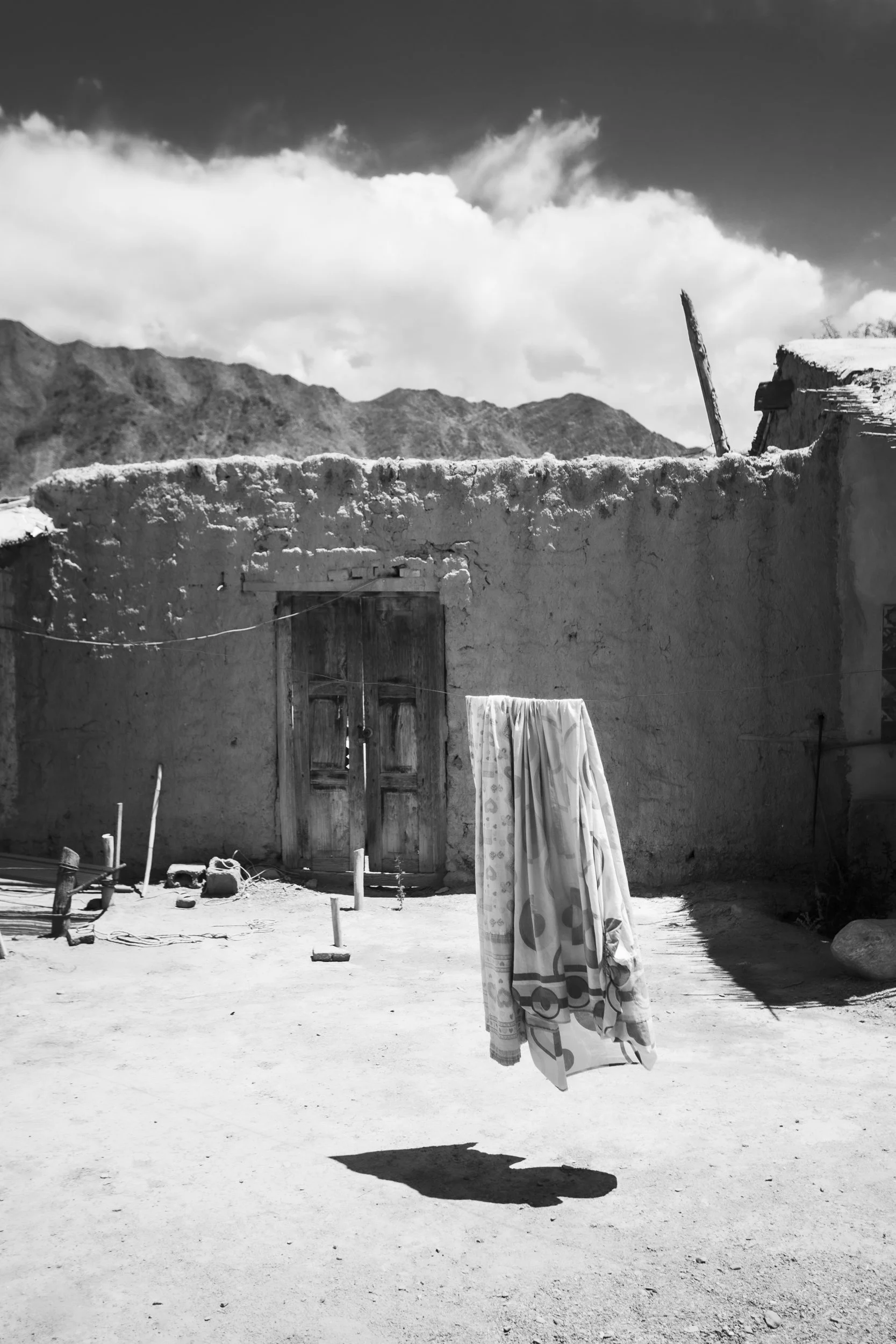 A black and white photo of a rural outdoor scene with a dirt ground, old mud wall building, clothes hanging on a line, and mountainous background under cloudy sky.