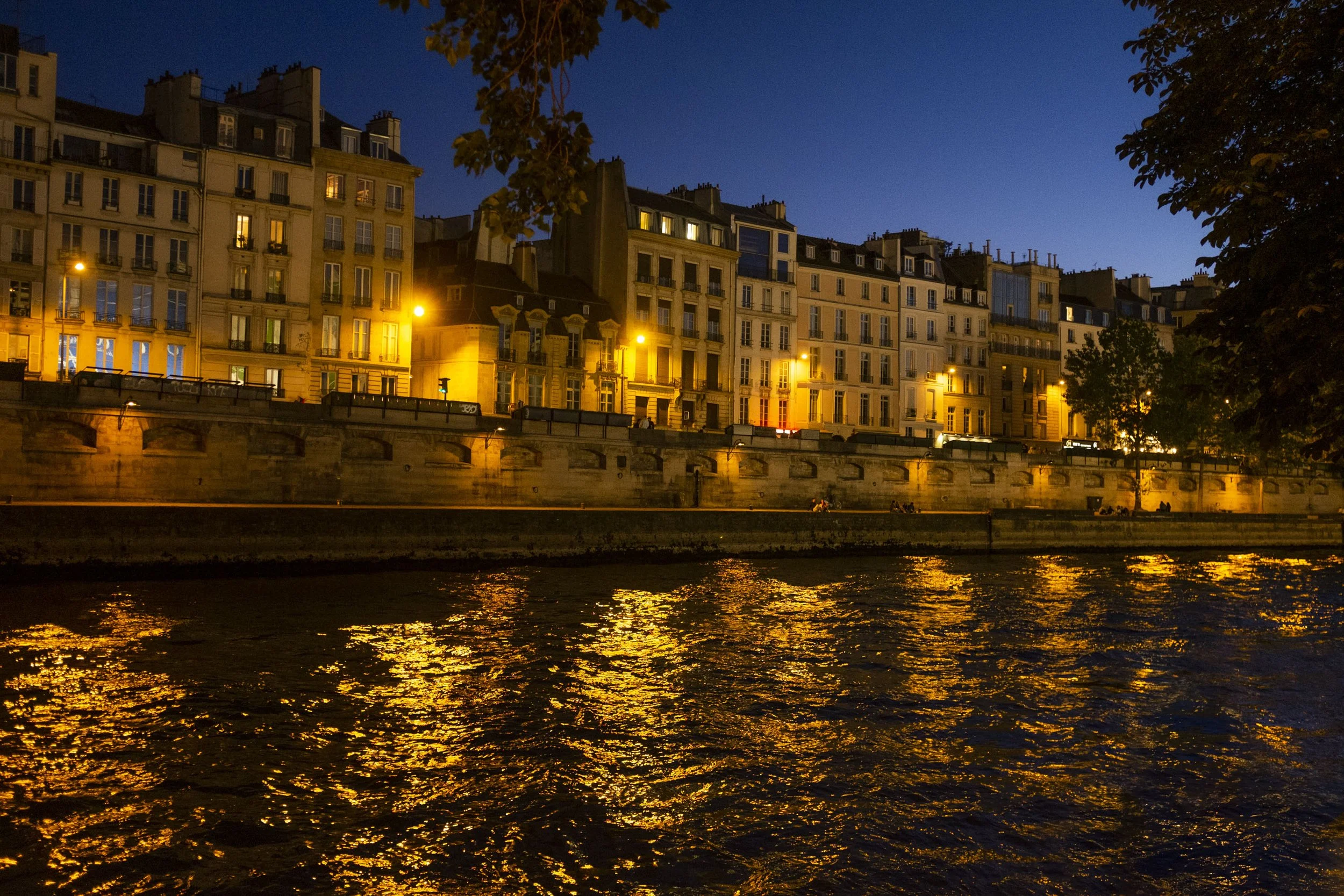 Cityscape of buildings along a river at dusk, with warm yellow streetlights and dark blue sky reflected in the water.