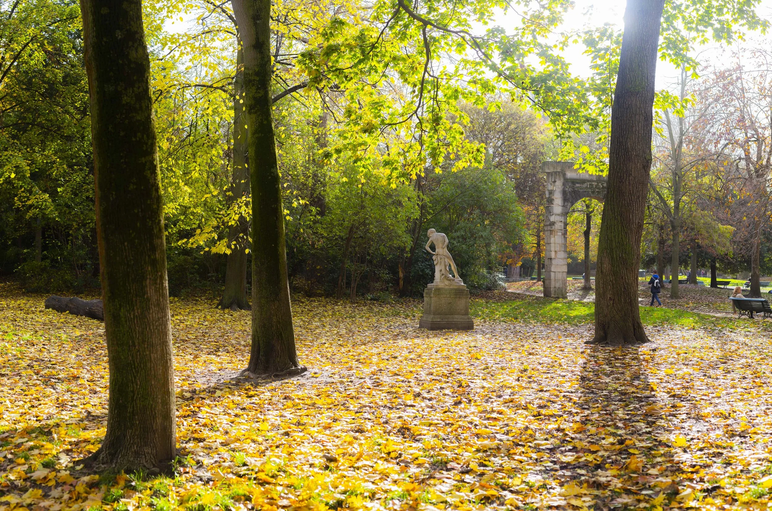 A park scene in autumn with trees, fallen yellow leaves on the ground, a white statue of a woman, and columns in the background. There are benches and people walking in the distance.