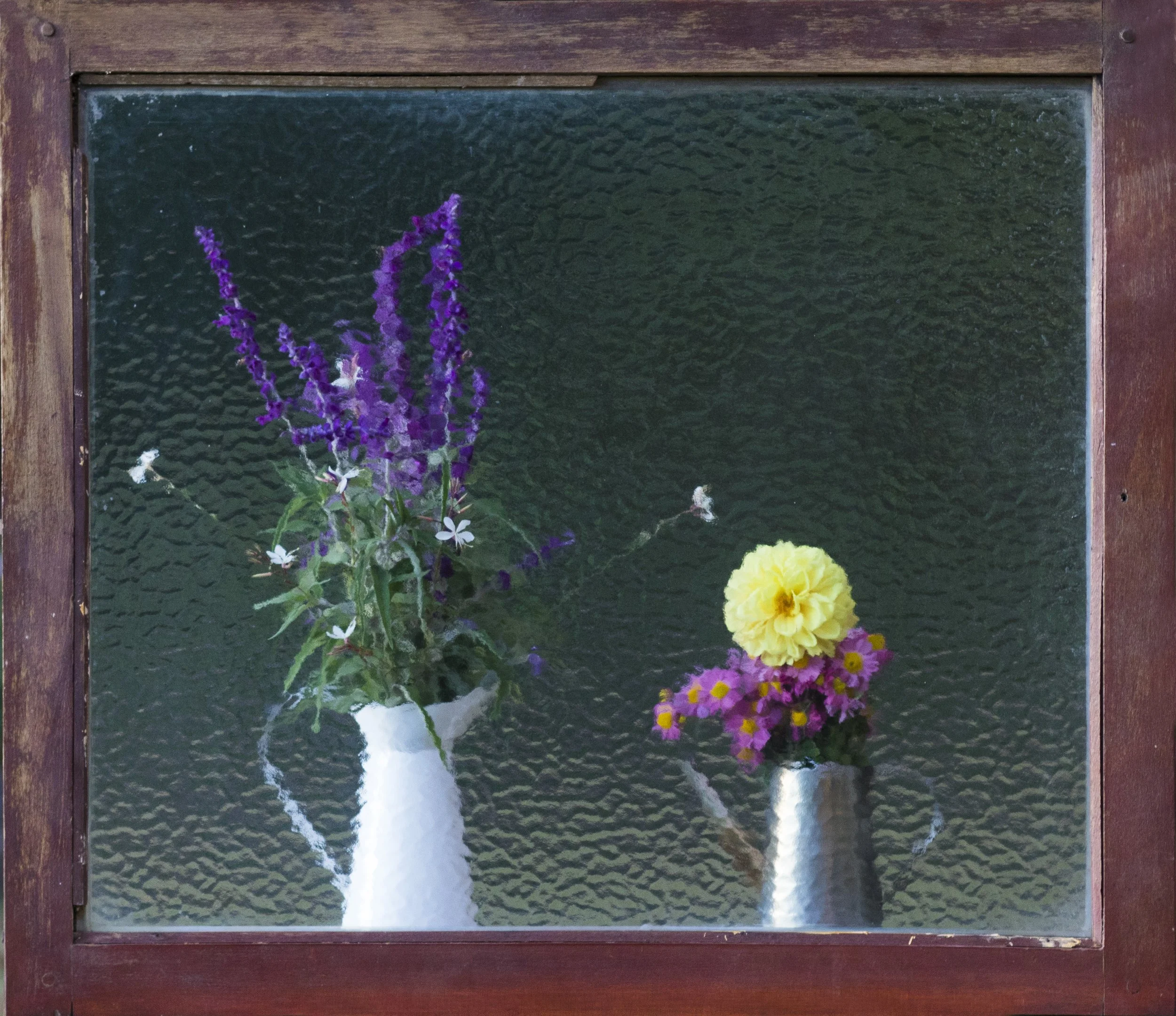A window with water and blurry reflections, a purple flower with elongated petals, and a yellow flower with ruffled petals visible in the reflection.