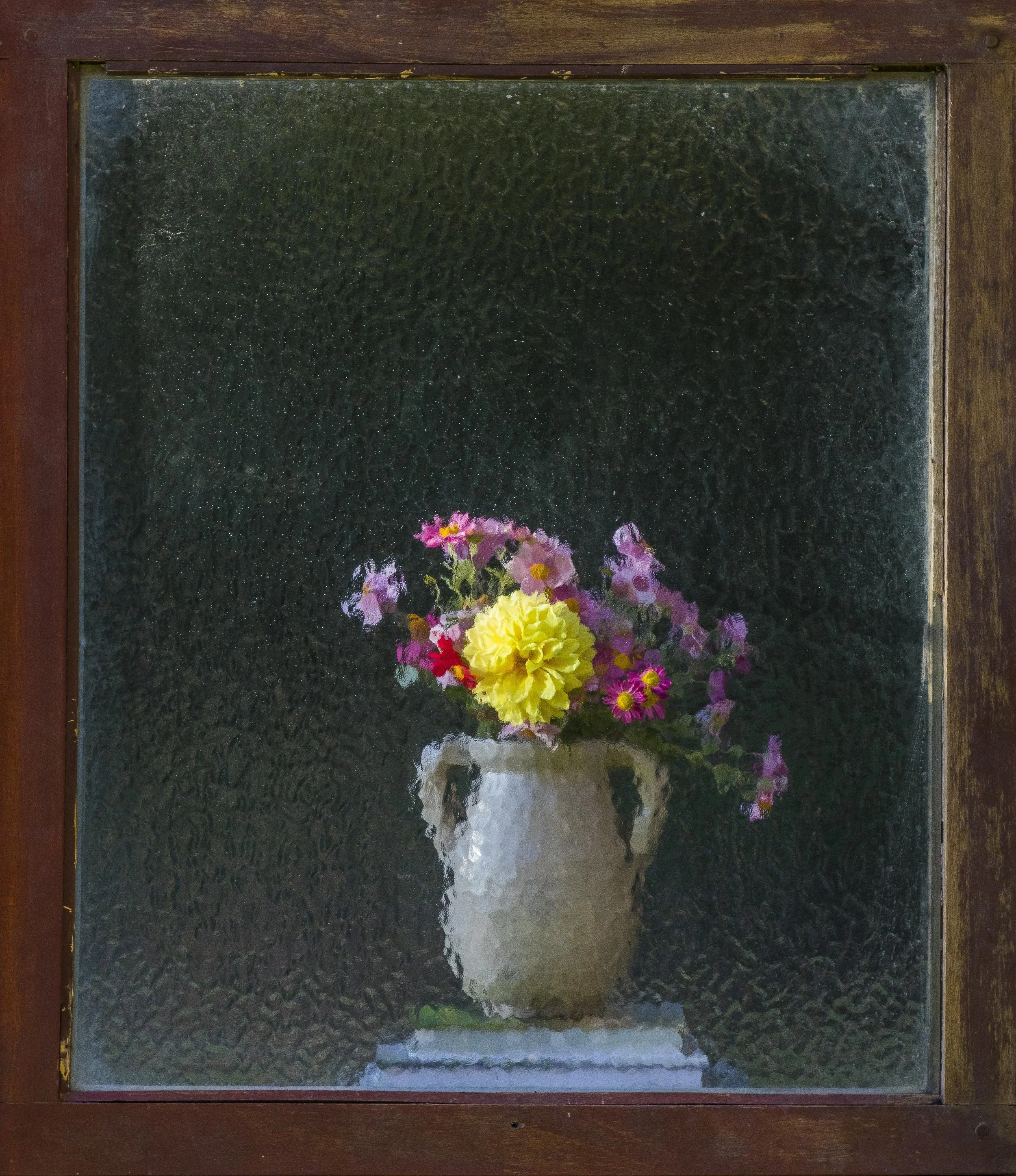 Framed mirror reflecting a bouquet of pink, yellow, and purple flowers in a vase. The mirror is mounted on a wooden surface.