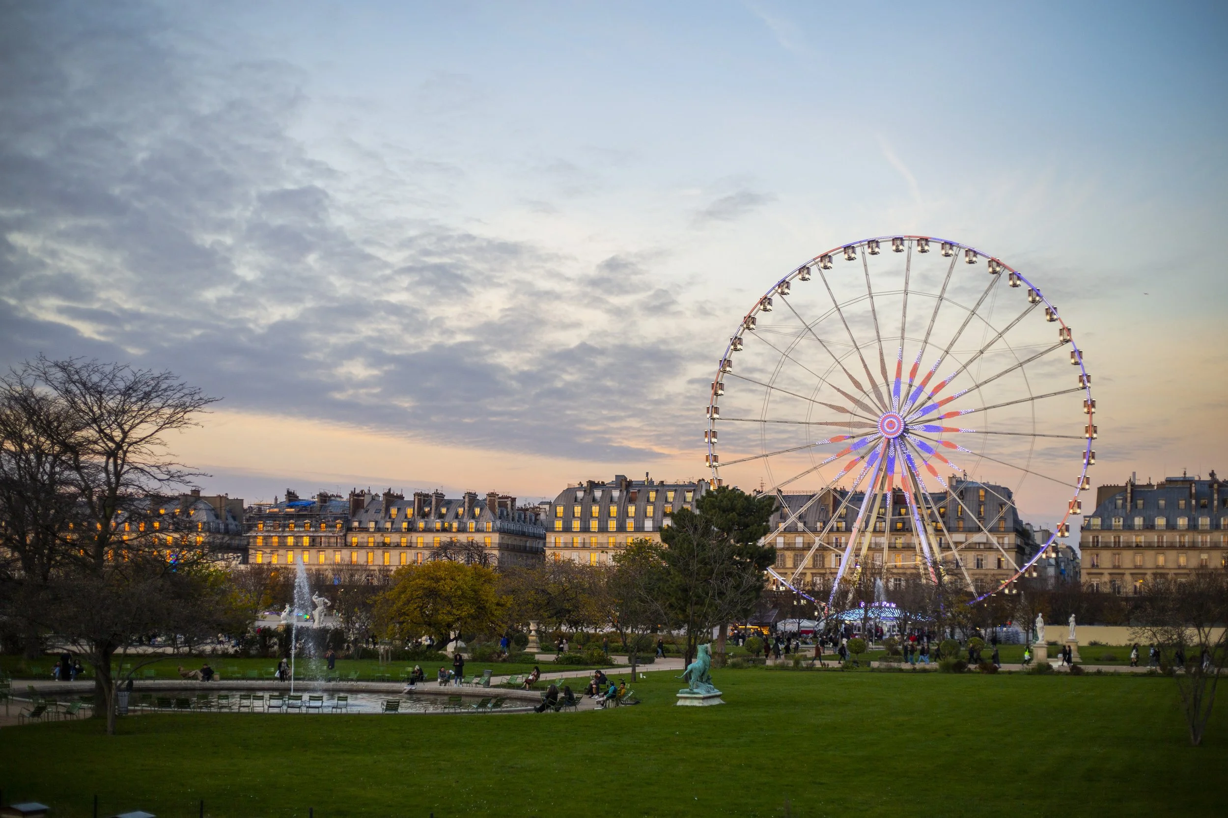 Paris city park at dusk with a large illuminated Ferris wheel on the right, historic buildings with lit windows in the background, a fountain with statues in the foreground, and people walking and sitting around the park.