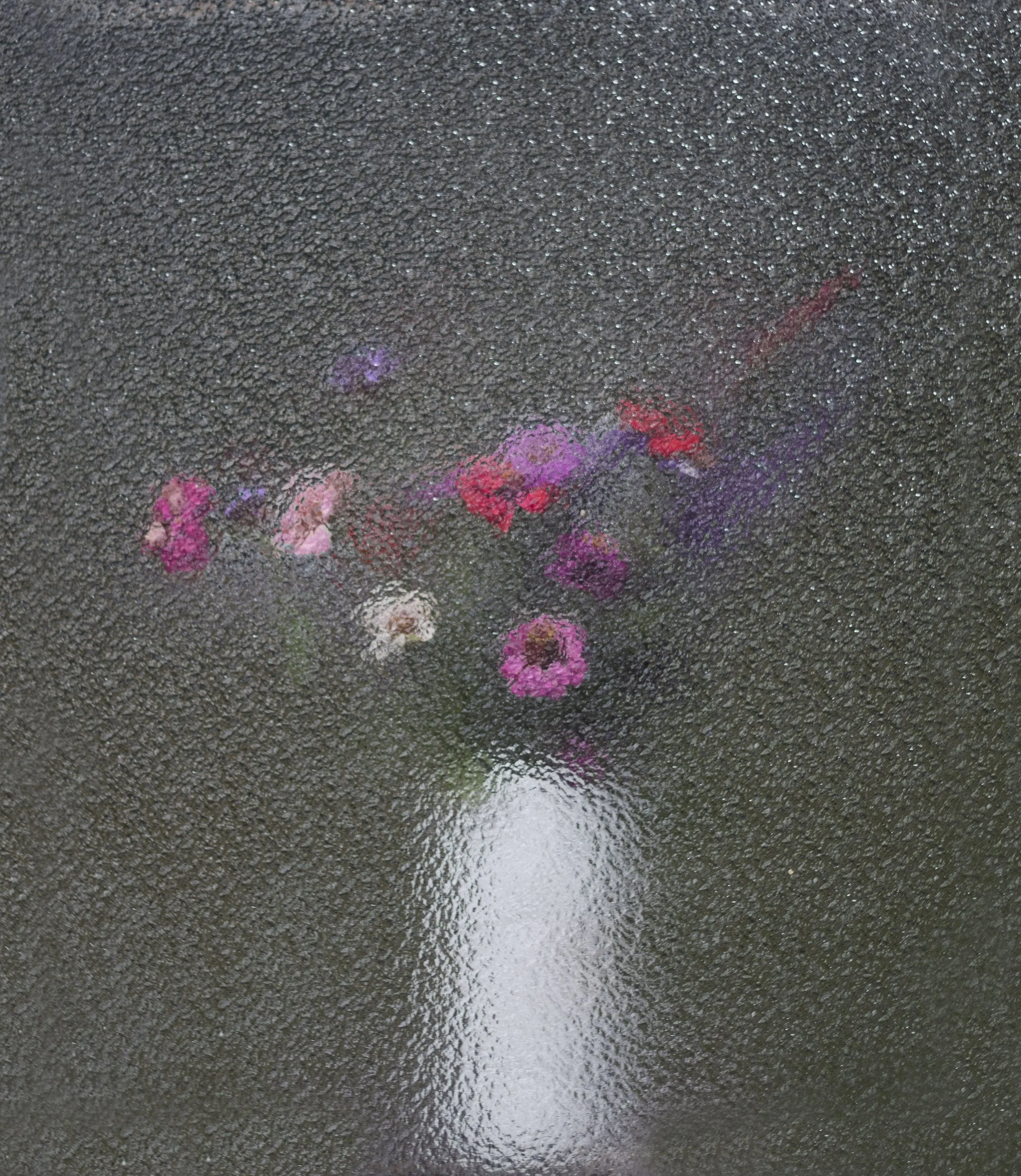 A wet, shiny, textured surface, possibly a dark asphalt or concrete sidewalk, with a reflection of colorful flowers or objects, mainly pink, purple, and red, slightly blurry on the lower part of the surface.