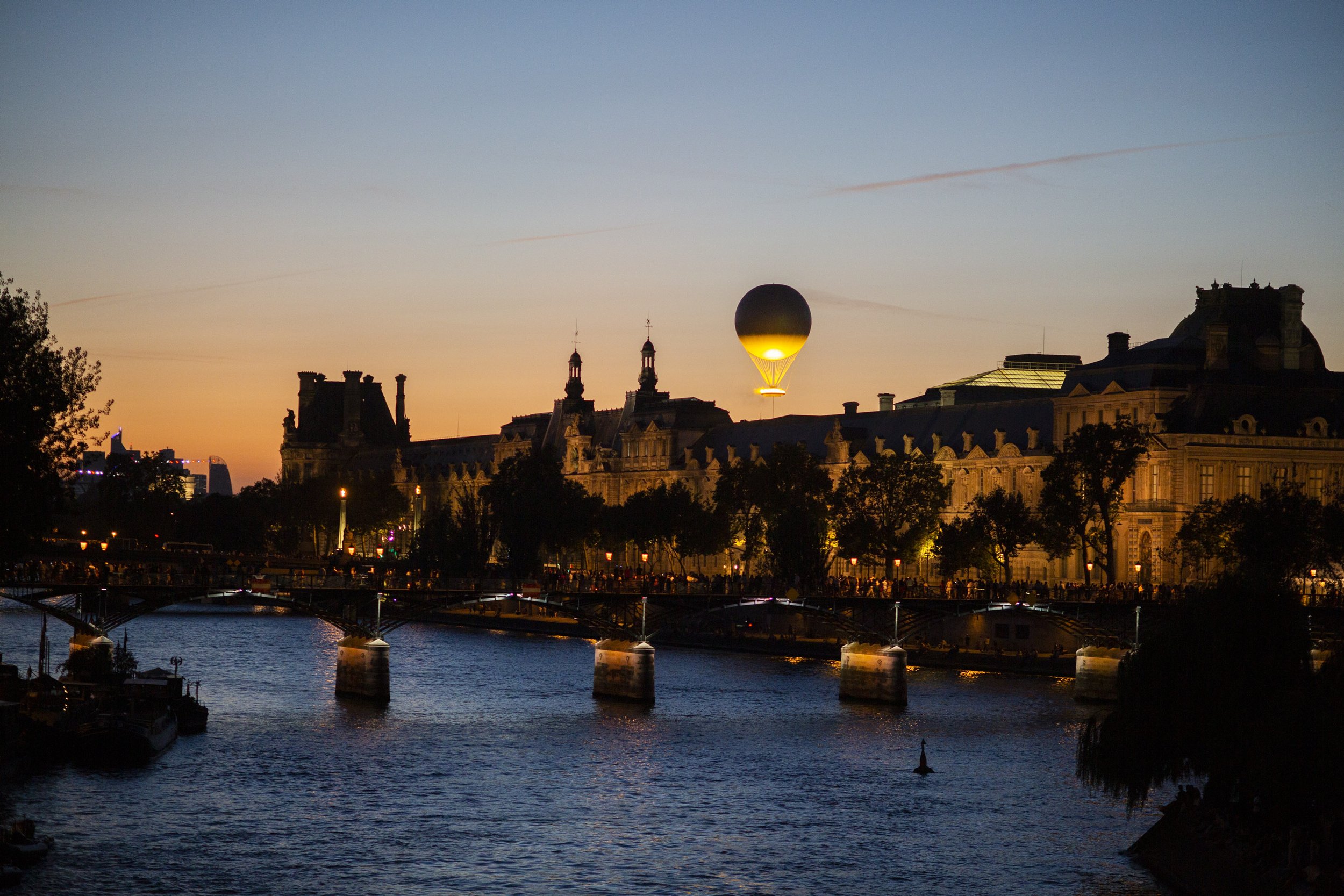 A cityscape during sunset with a hot air balloon in the sky over historic buildings and a river in the foreground.
