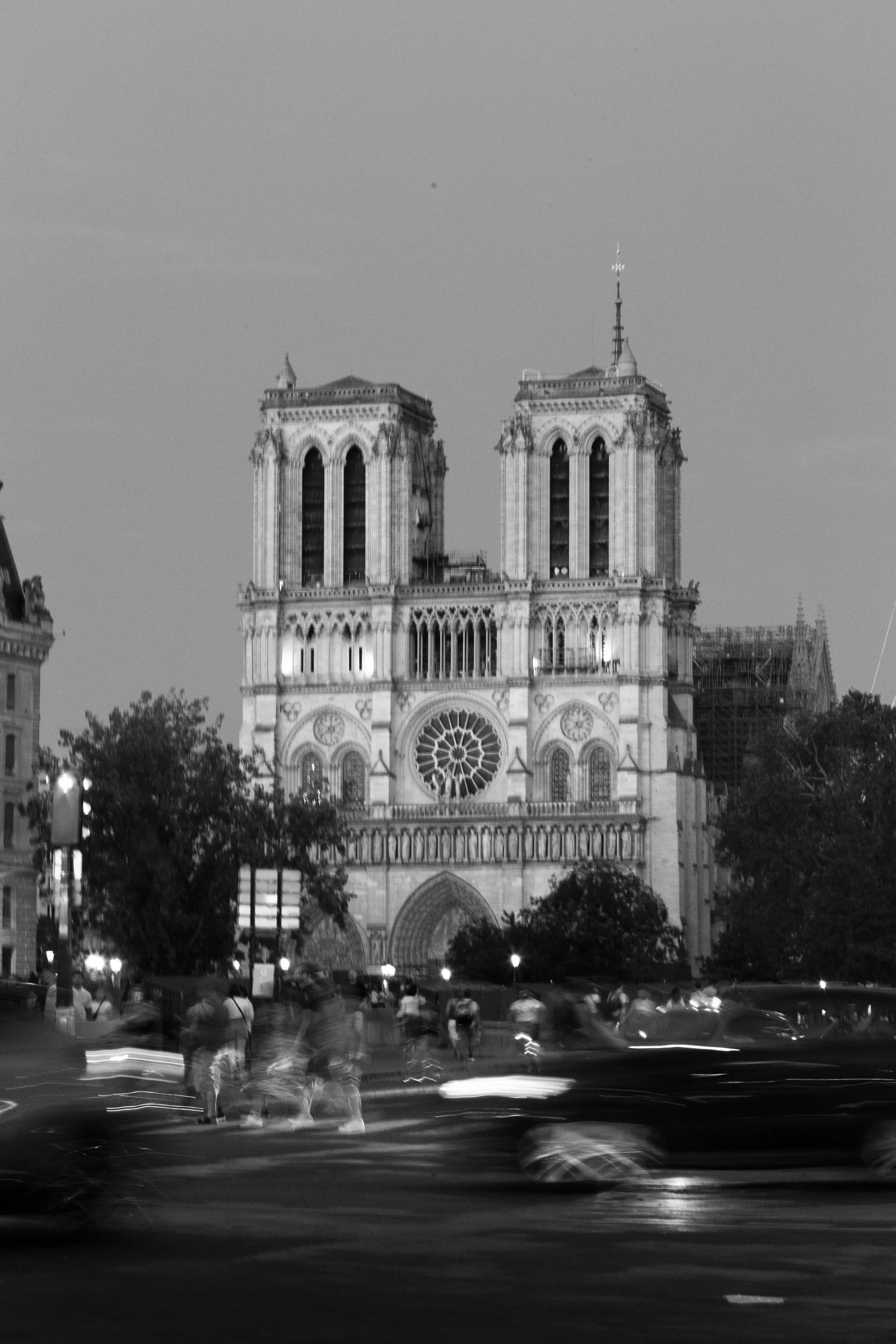 Nighttime view of Notre Dame Cathedral in Paris, France, with blurred cars and people in the foreground.