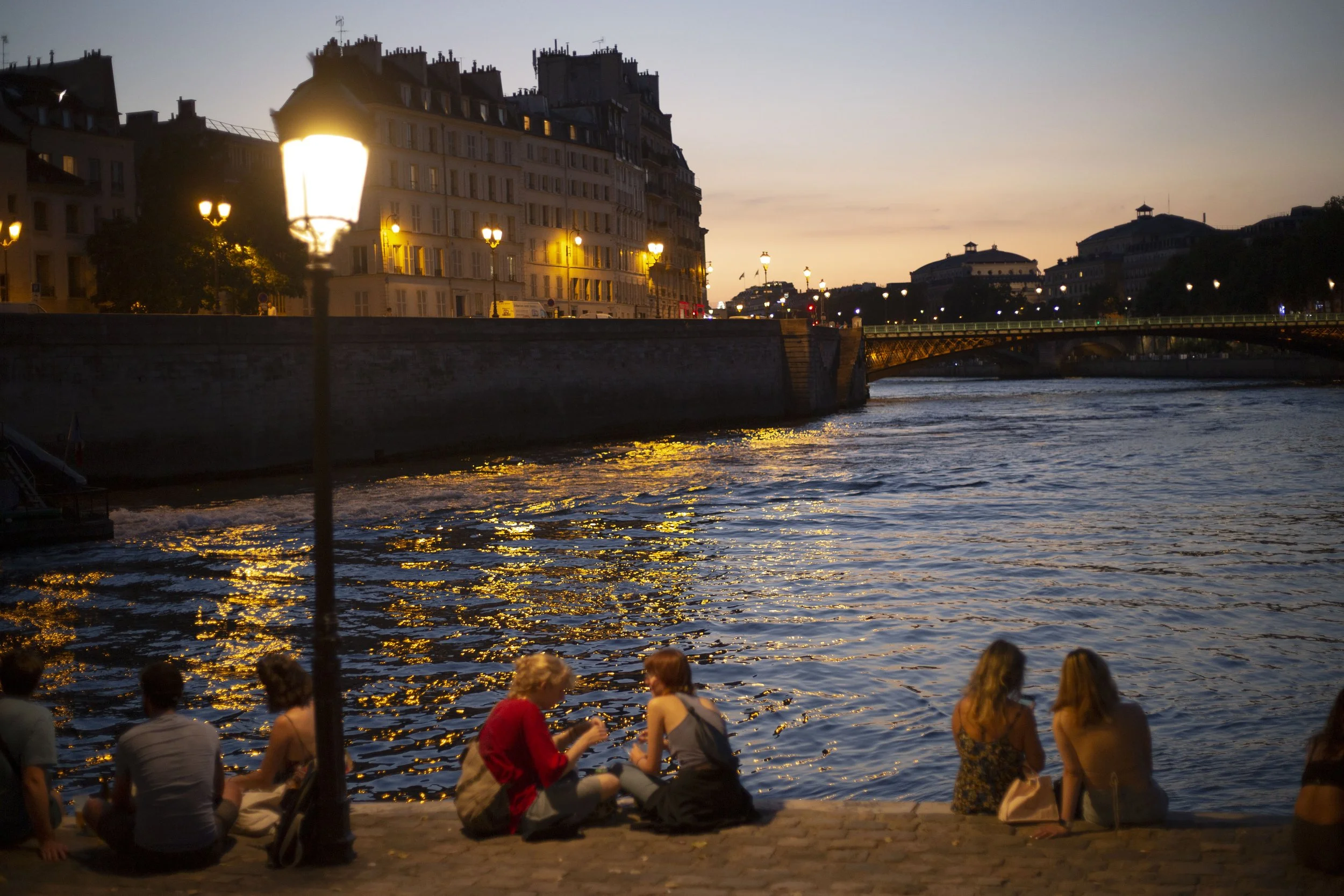 People sitting by the riverbank watching the sunset with historic European buildings and a bridge in the background.