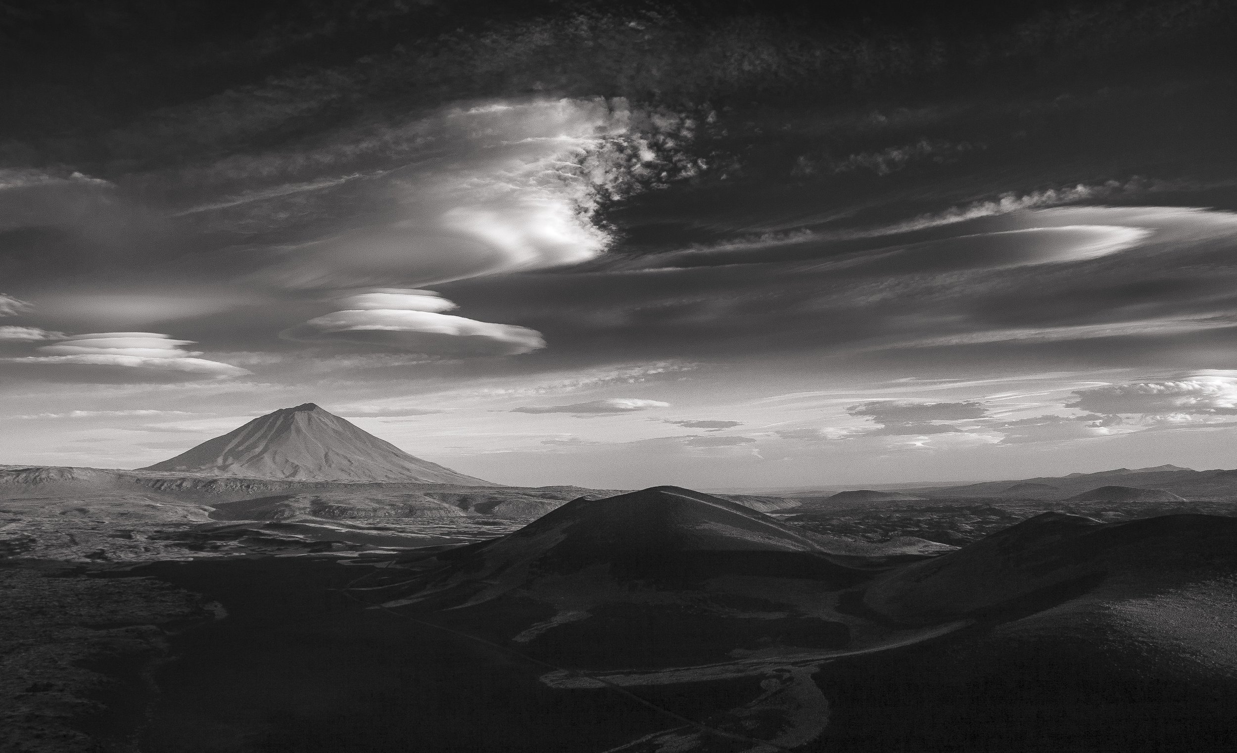 A black and white landscape featuring a large mountain with a conical shape and a flat top, set against a dramatic sky with swirling clouds.
