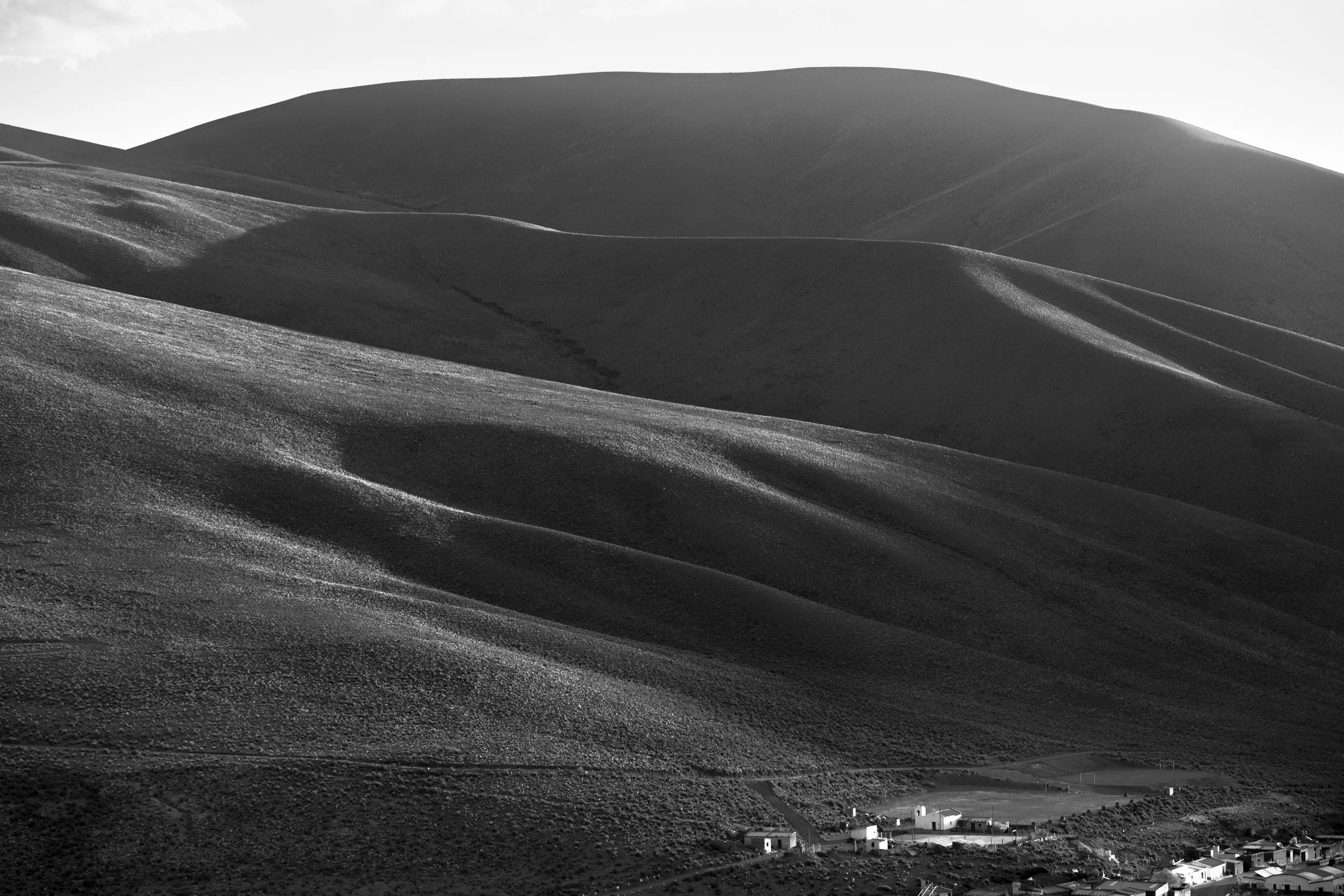 Black and white photo of rolling hills with a small village at the foot of the hills. The landscape is illuminated by sunlight, creating shadows on the hills.